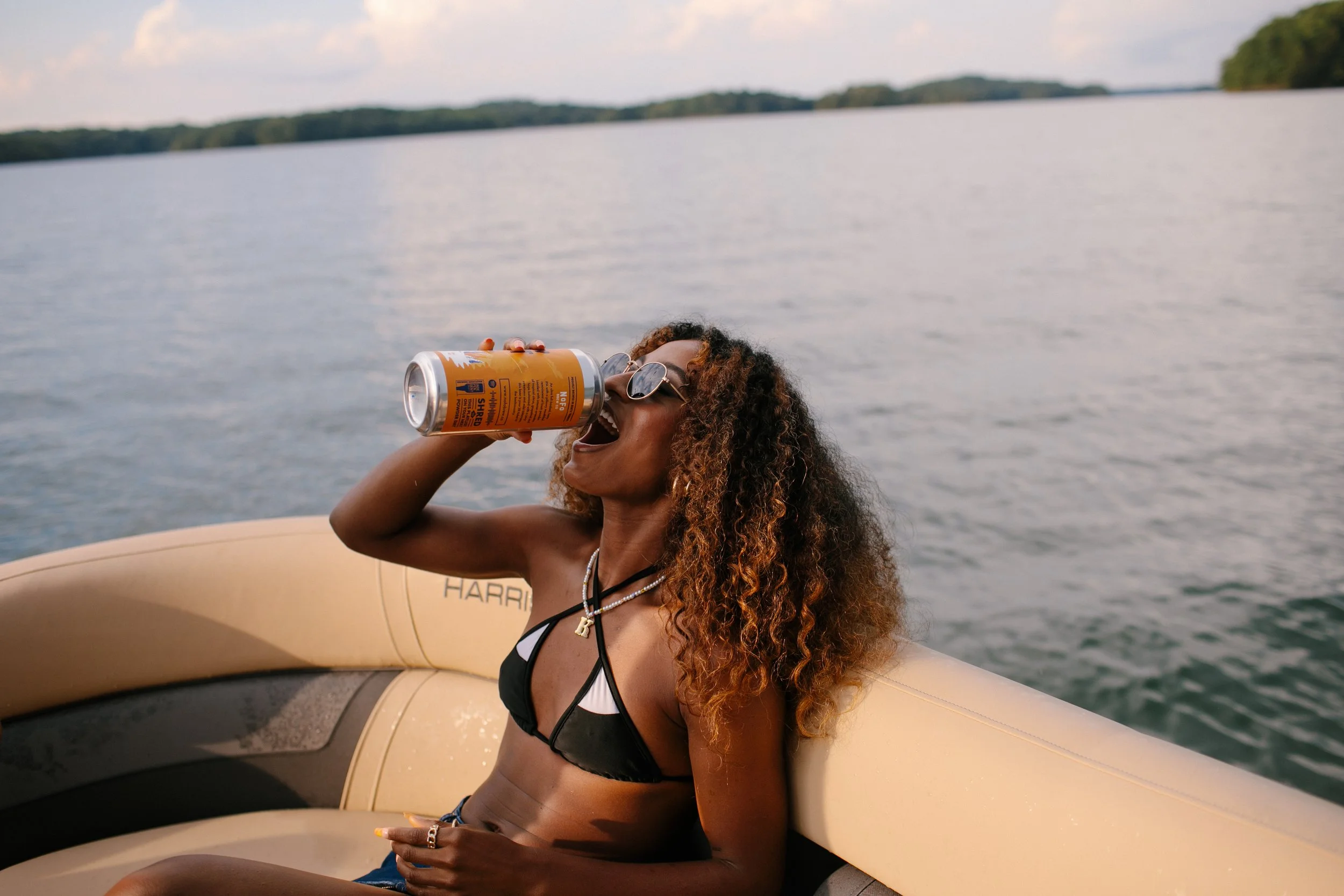 A woman with curly hair and sunglasses enjoying a drink from a can while sitting in a boat on a lake.