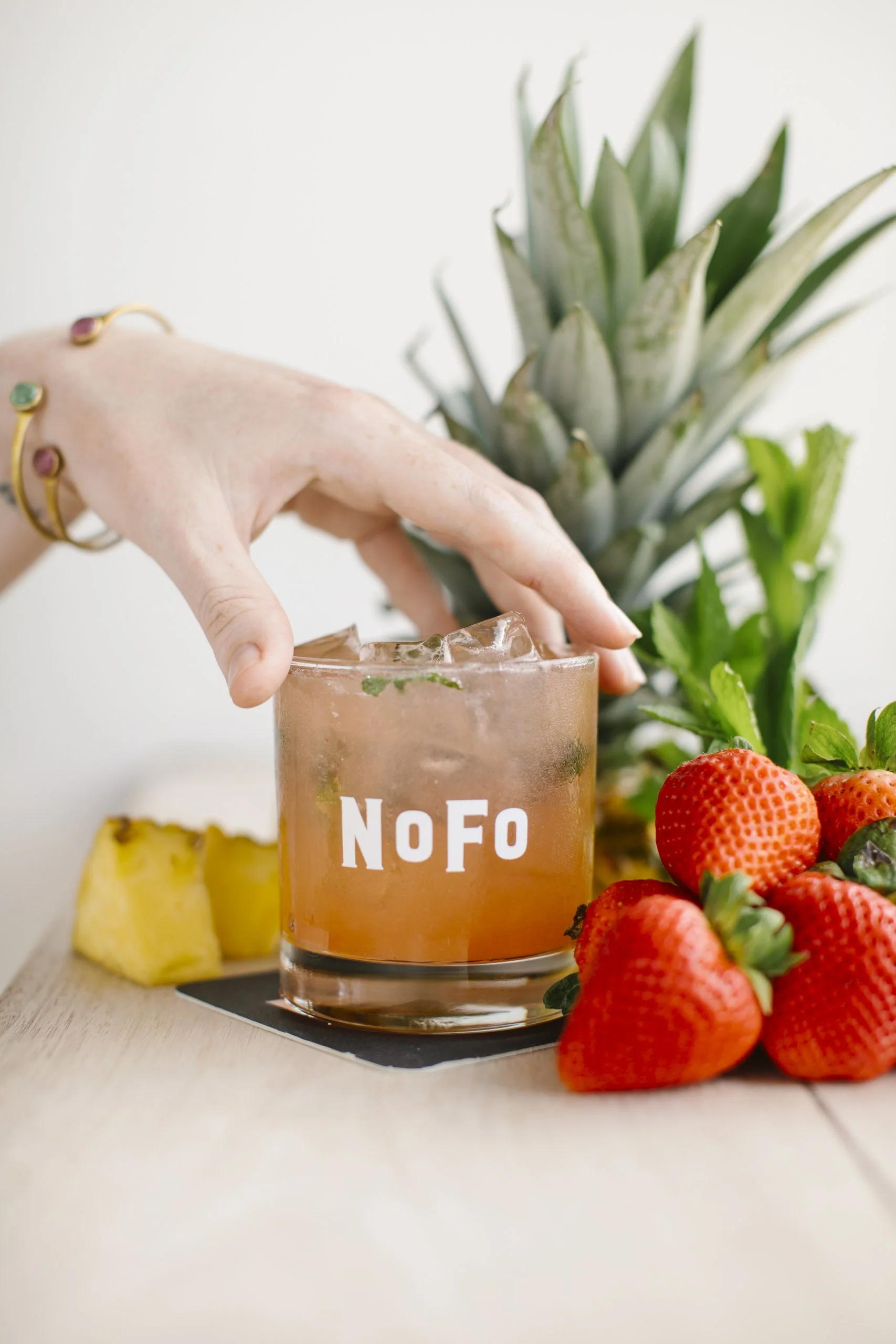 A hand with gold jewelry reaching for a pink drink in a glass labeled 'NoFo', surrounded by fresh strawberries, pineapple chunks, and a pineapple in the background.