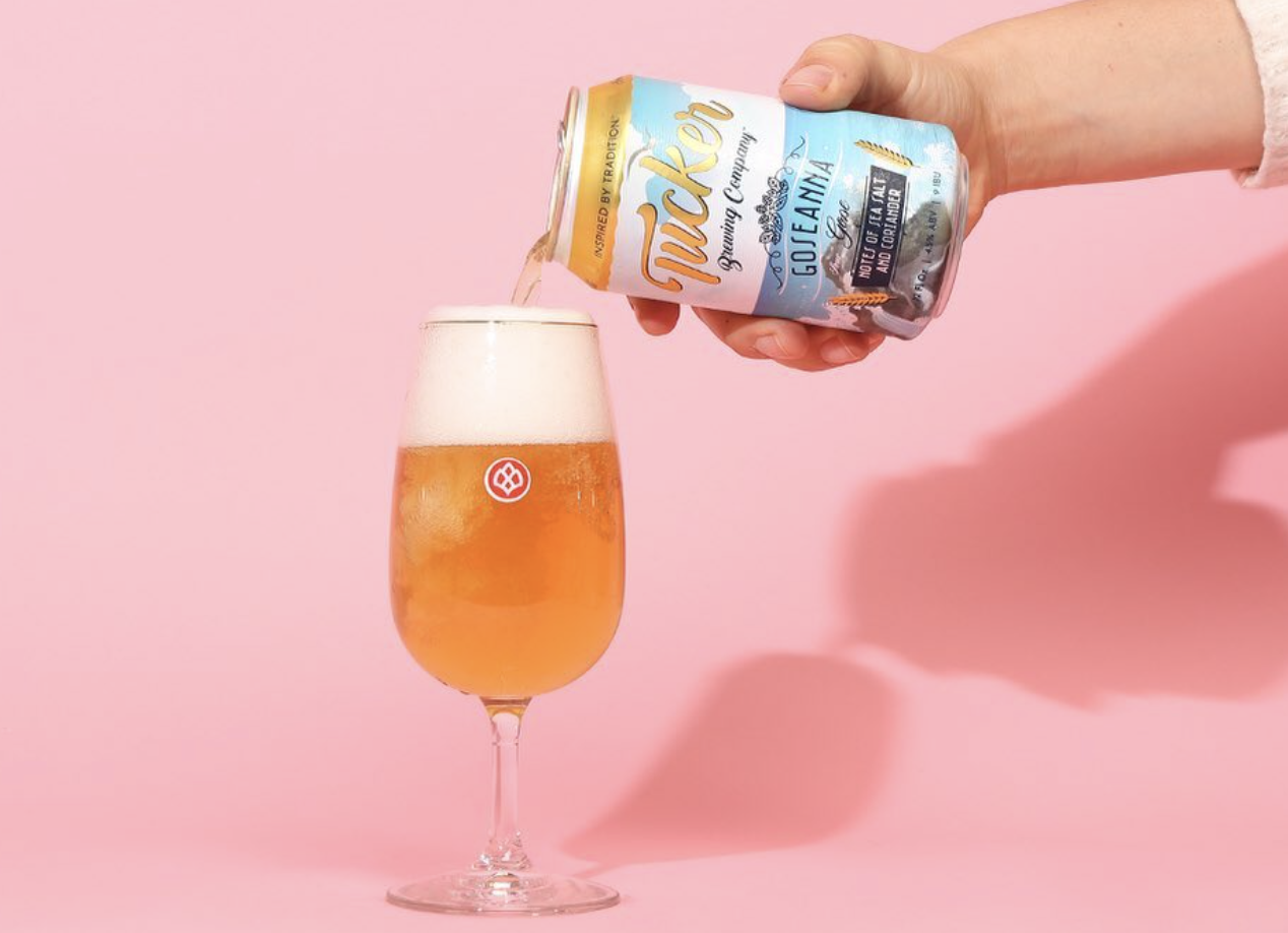 A hand pouring a can of beer into a tall glass filled with beer against a pink background.
