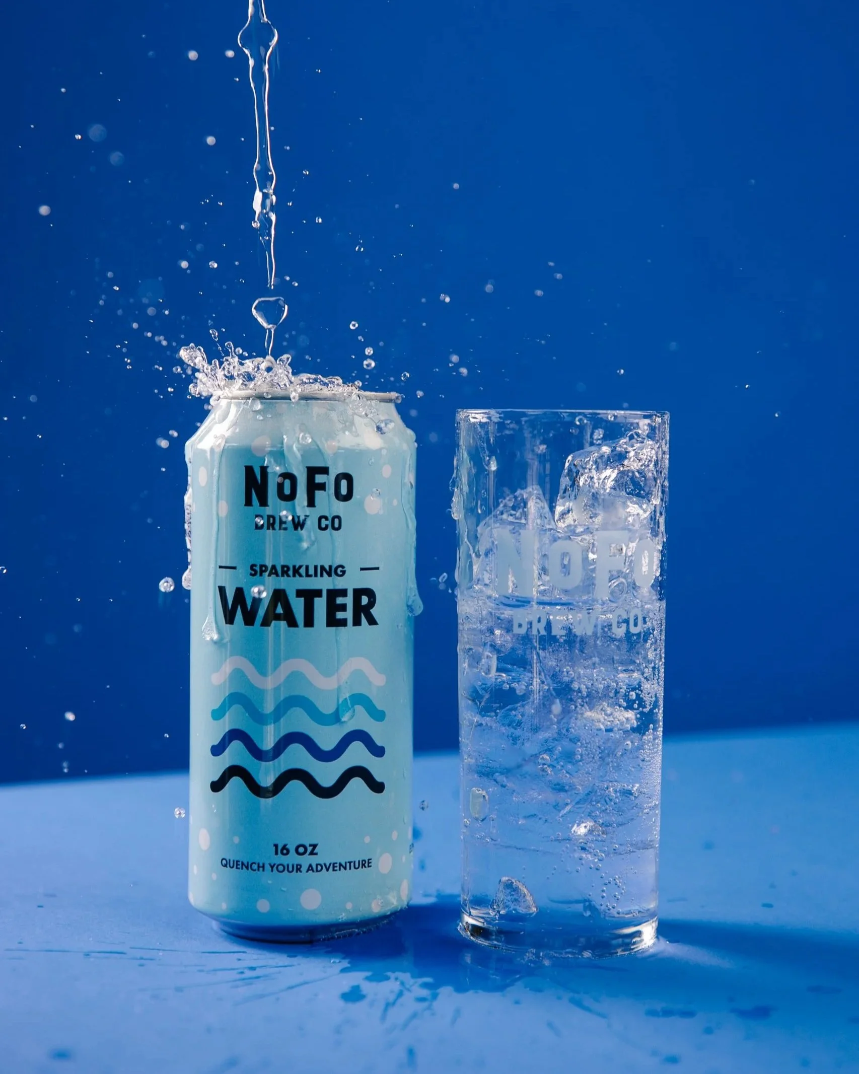 A can of sparkling water with bubbles appears to be overflowing into a glass that is filled with ice, with the ice and bubbles spilling over the edges. The background is a solid blue.