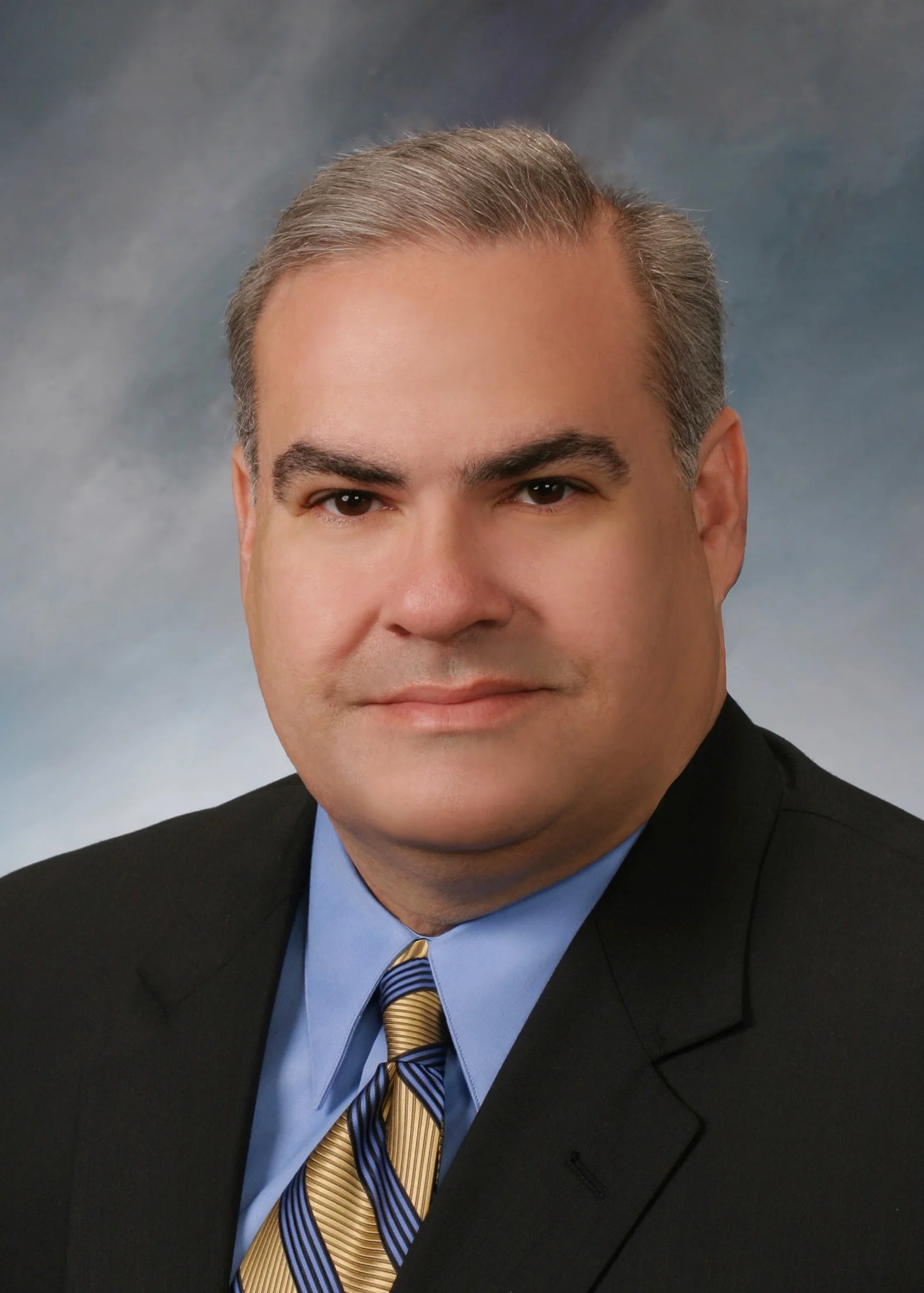 A professional headshot of a man in a suit and tie, with gray hair, against a blue-gray background.