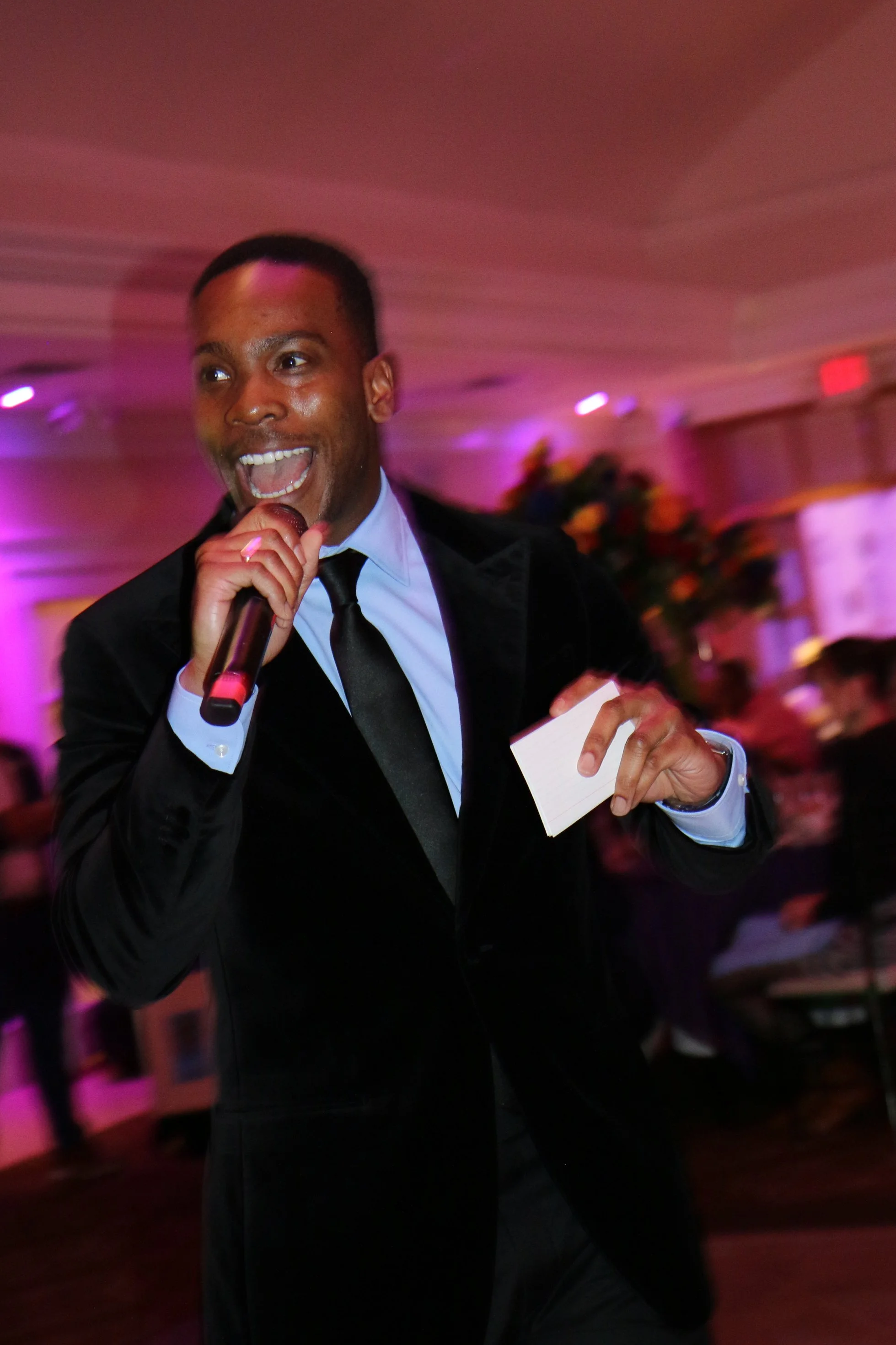 A man in a tuxedo singing into a microphone at an event or celebration with pink and purple lighting.