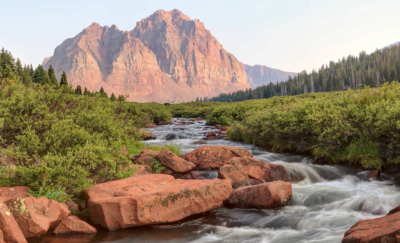A river flowing over rocks surrounded by green bushes, with large red sandstone mountains in the background under a clear sky. North Slope of the Unita Mountains