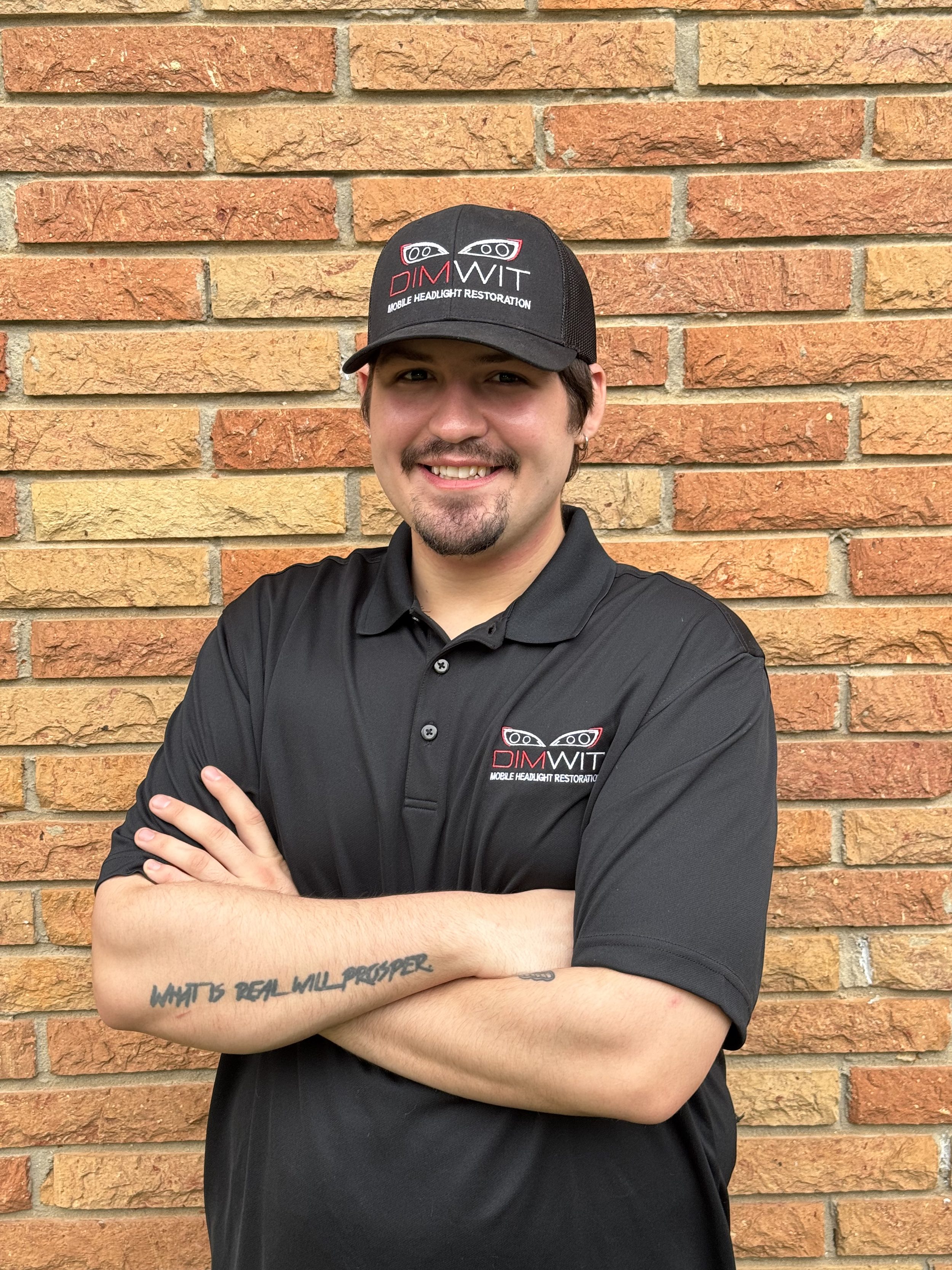 A young man with a goatee and mustache, wearing a black baseball cap with a logo and the words 'DIM WIT Mobile Headlight Restoration', stands in front of a brick wall.