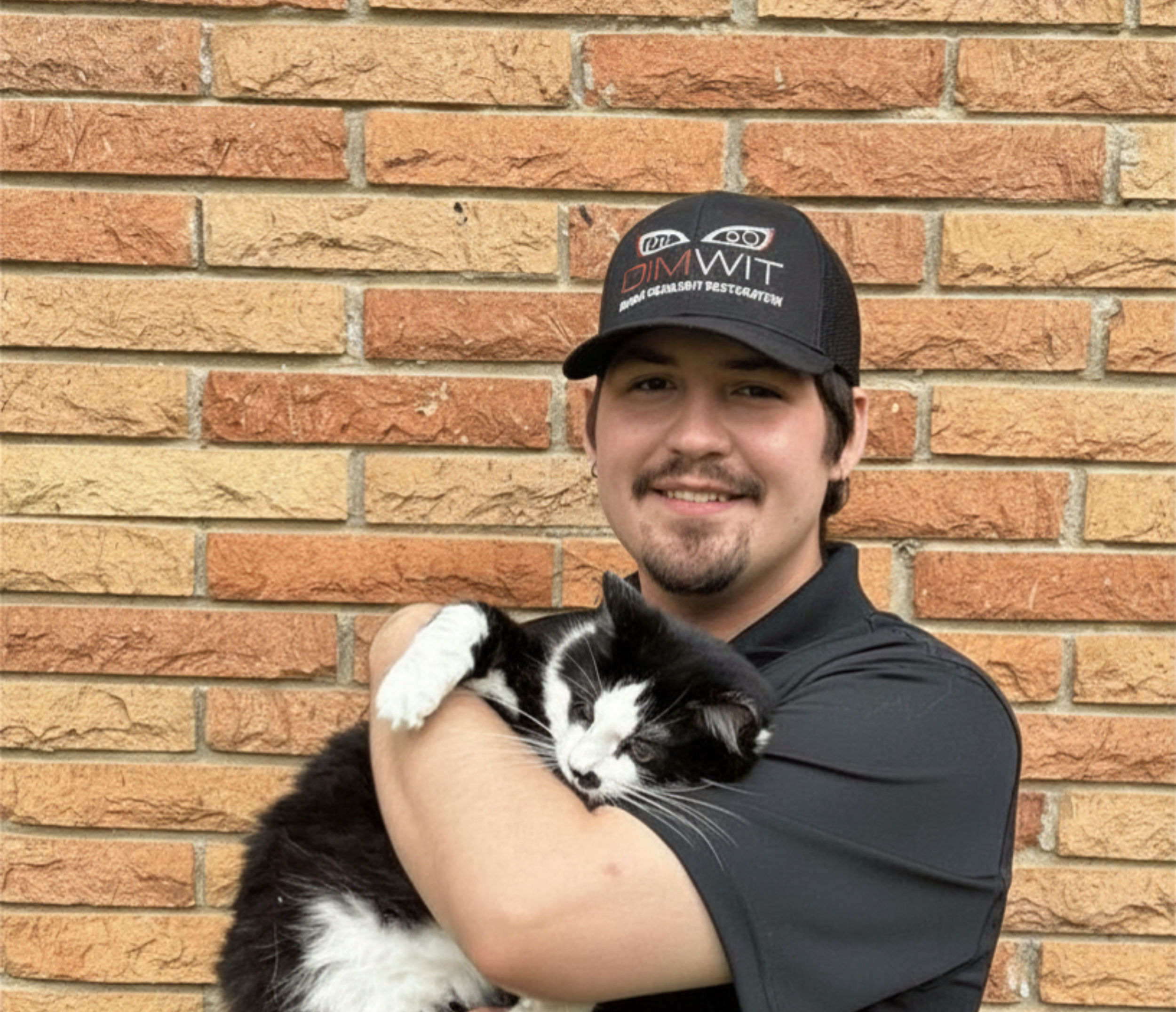 A man wearing a black cap and black shirt, holding a black and white kitten, standing against a brick wall.