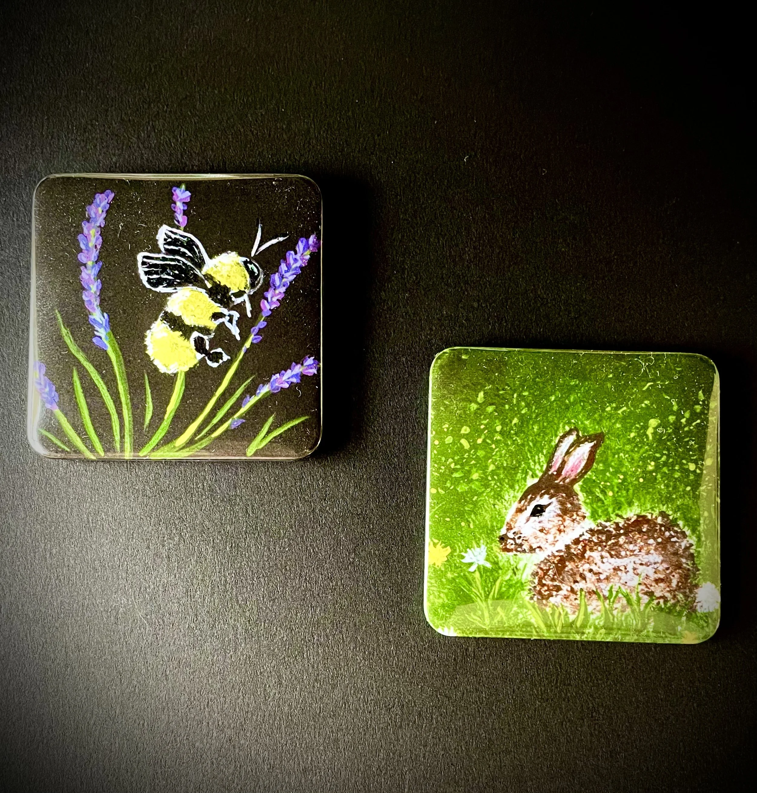 Close-up of two rectangular stickers with nature illustrations on a black surface. One sticker shows a bumblebee on yellow flowers with lavender stems; the other depicts a rabbit in a green grassy field.
