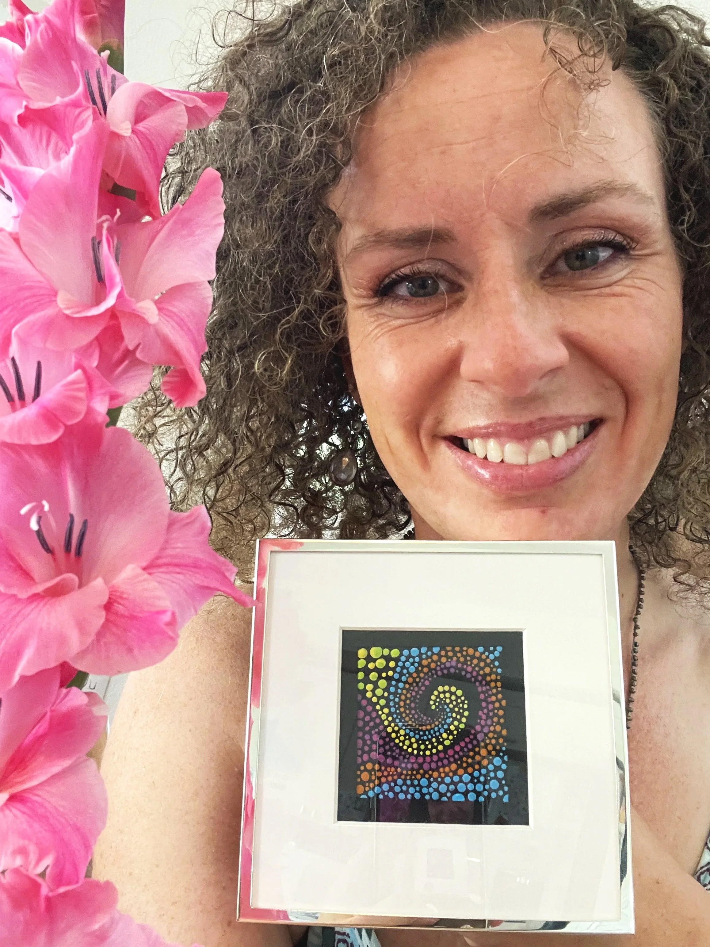 A smiling woman with curly hair holding a colorful dot-patterned artwork, with pink flowers in the foreground.