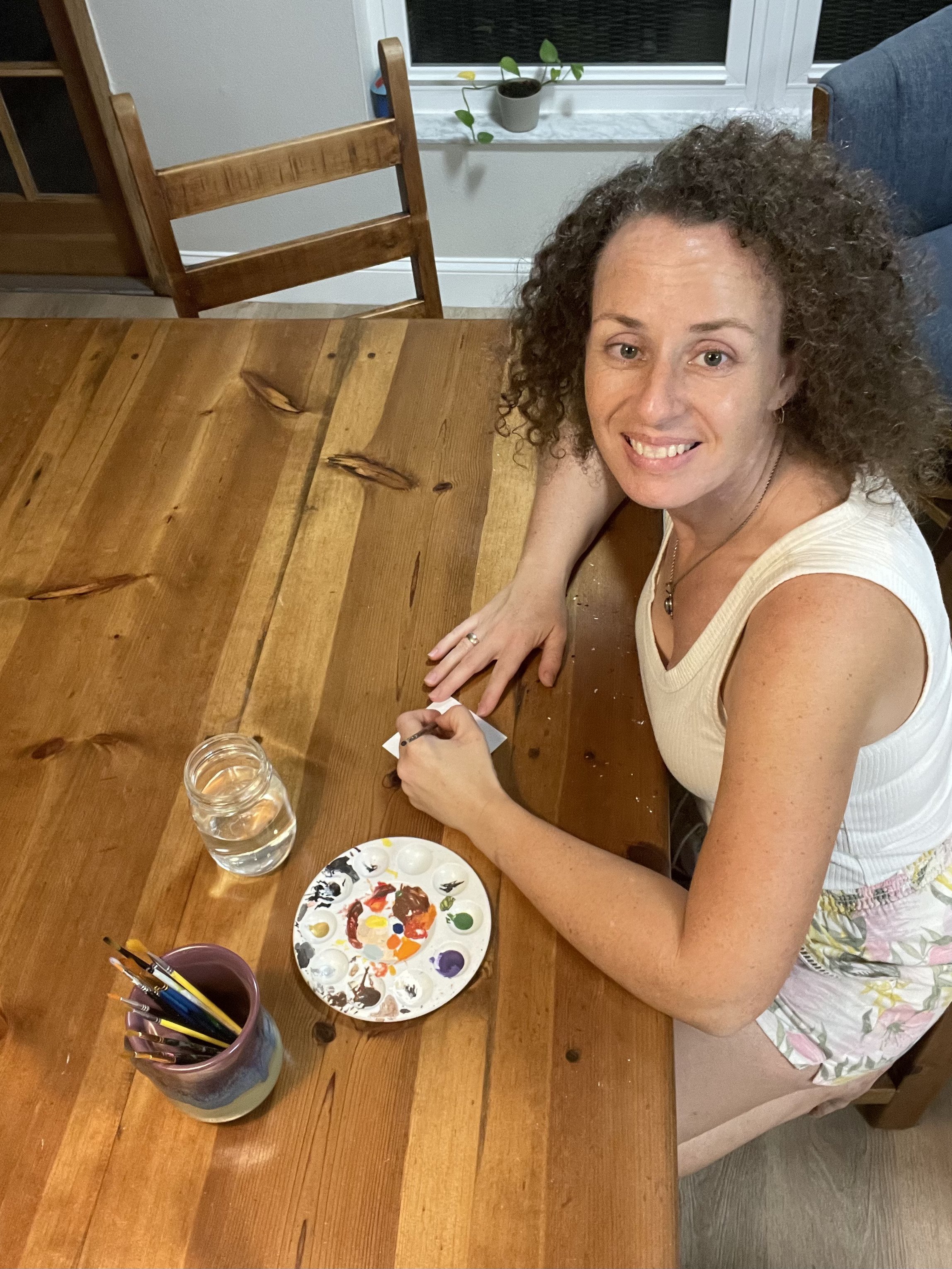 Woman with curly hair smiling at the camera, sitting at a wooden table with a paint palette, jars of water, and brushes, suggesting she is painting or having an art activity in a home setting.