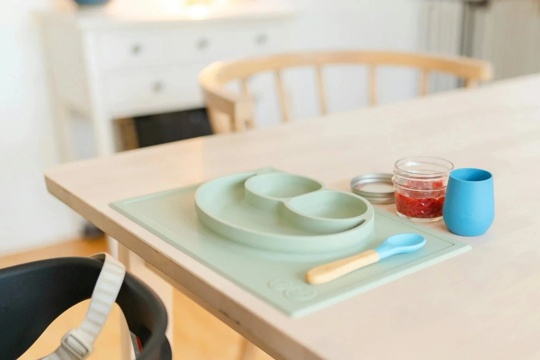 A children's breakfast set on a white table, including a mint green divided plate, a small blue cup, a glass jar with red jam, and a light-colored spoon. Part of a black high chair is visible in the foreground.