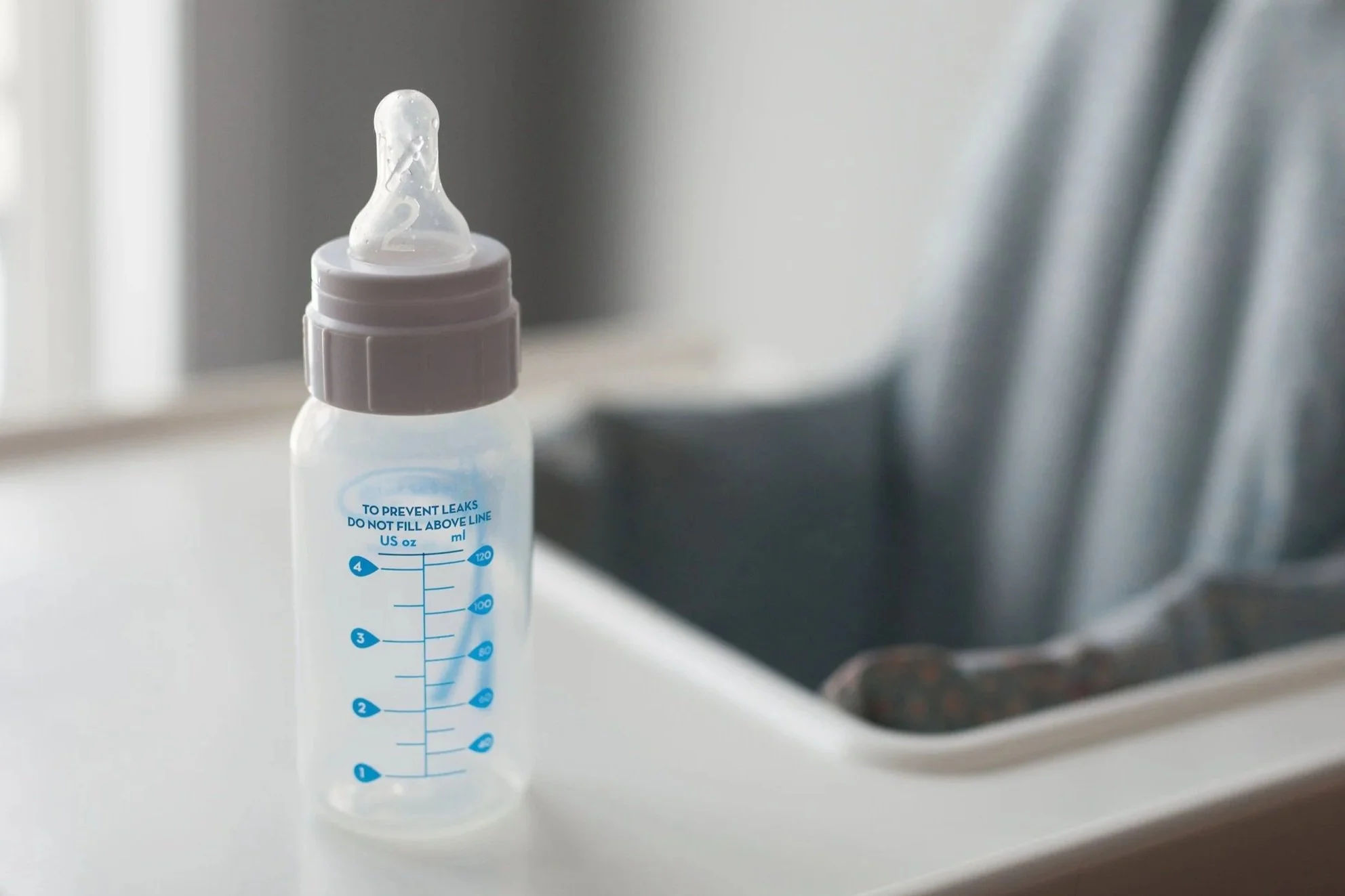 A baby bottle with a silicone nipple, placed on a high chair. The bottle has measurement markings.