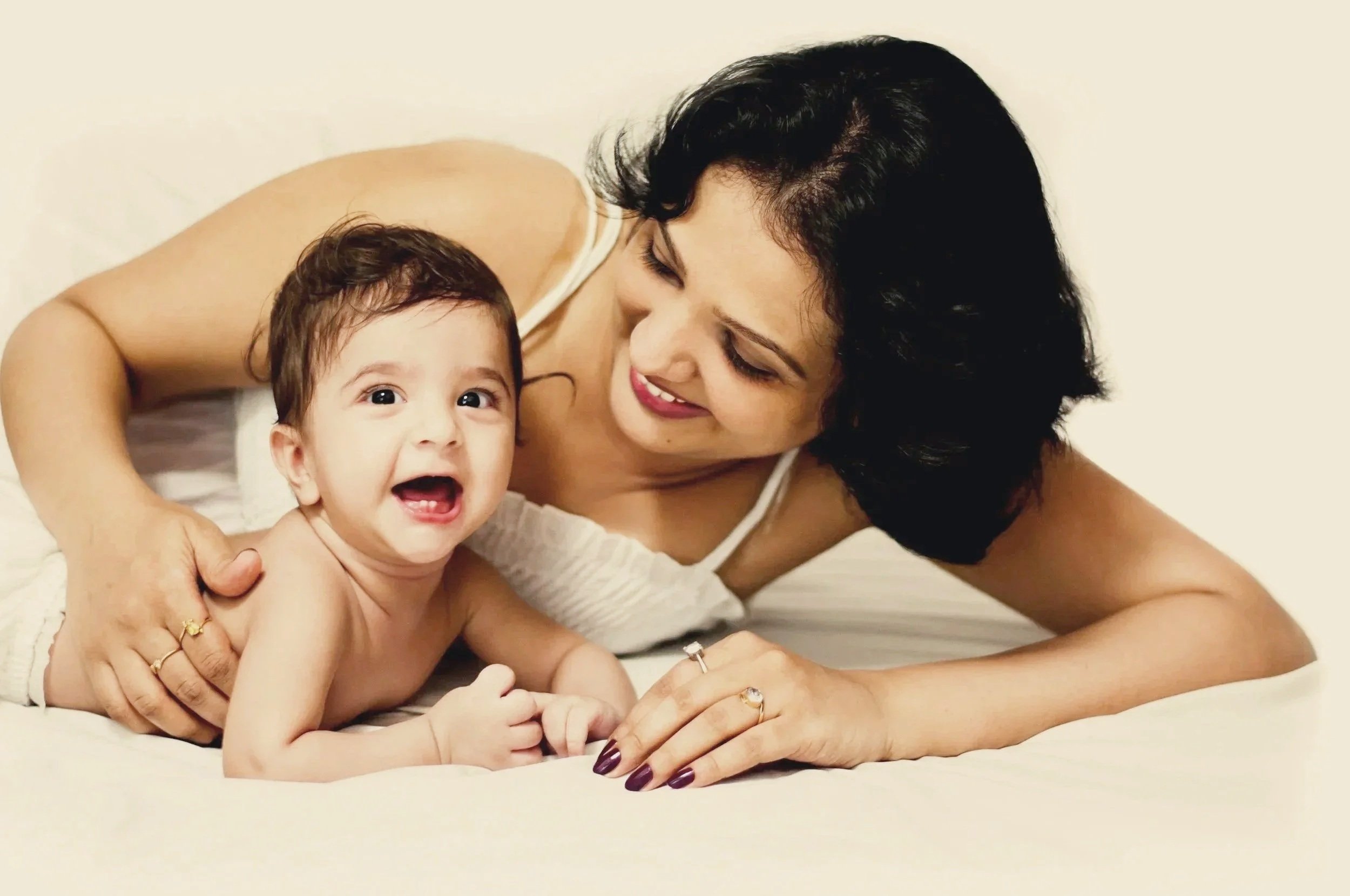 A woman and a young child lying on their stomachs on a bed, smiling and looking at each other, with a plain light-colored background.