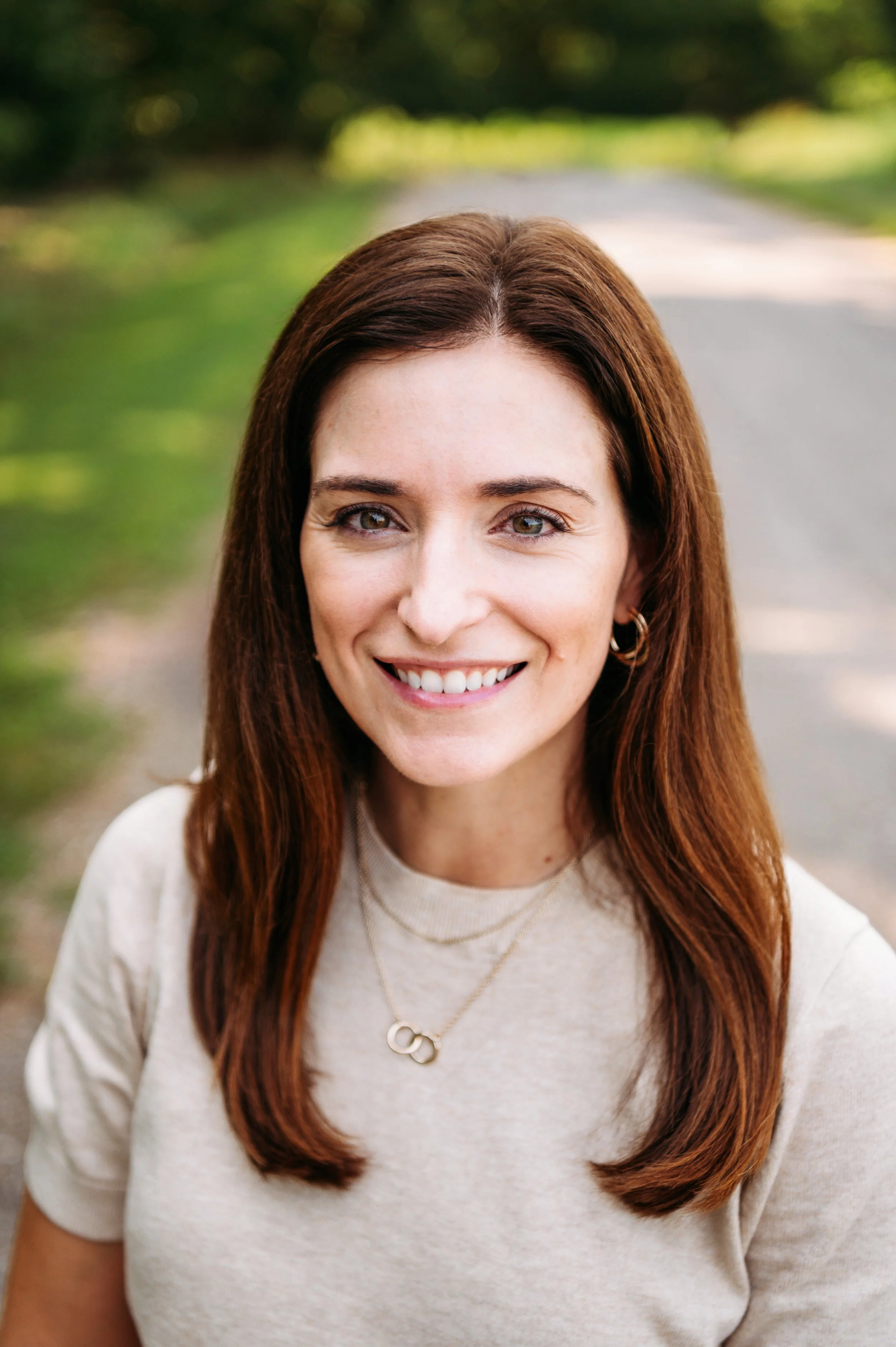 A woman with long brown hair and hazel eyes smiling outdoors on a pathway surrounded by greenery.