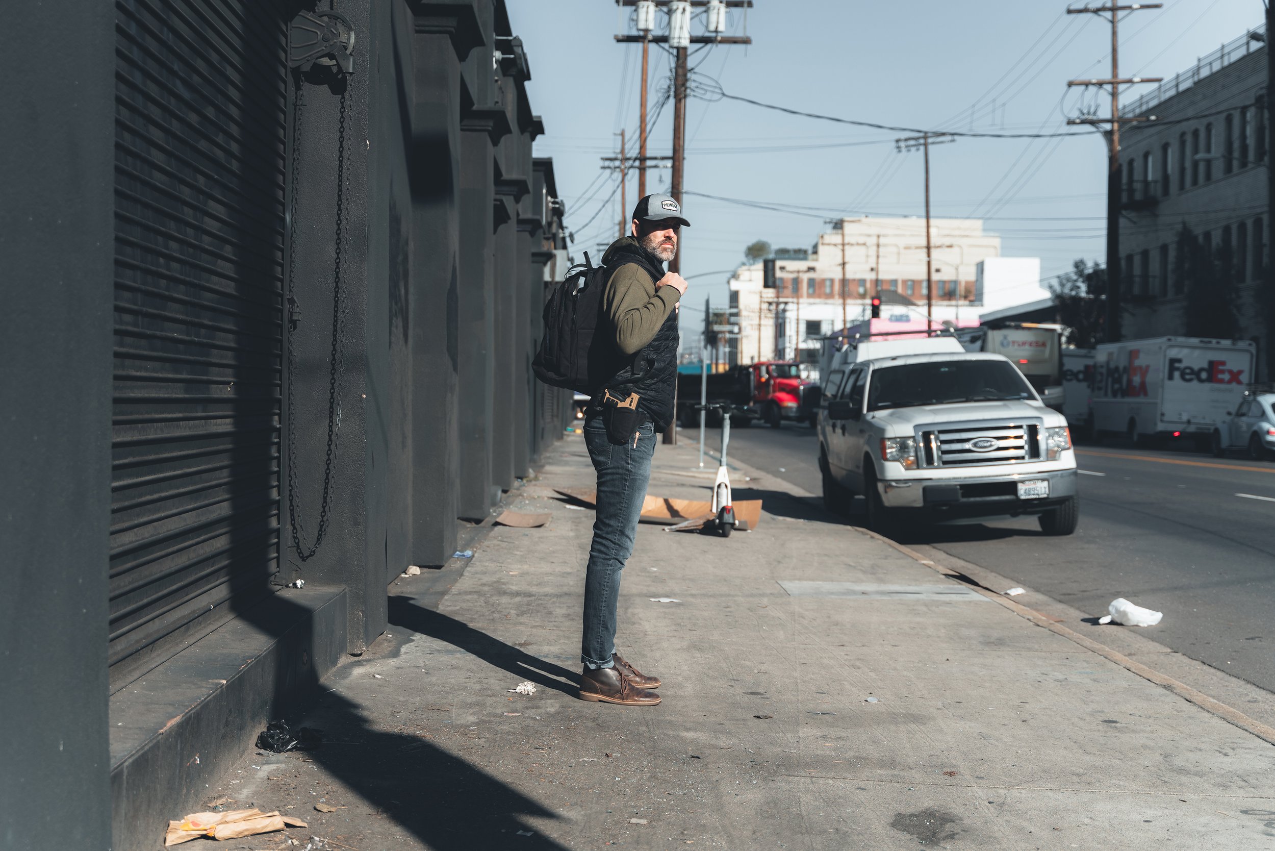 A man with a beard, sunglasses, and a cap, wearing a dark jacket and jeans, stands on a city sidewalk carrying a backpack. There are power lines and several parked vehicles, including FedEx trucks, on the street. The building behind him is dark-colored with a large vent or grille.