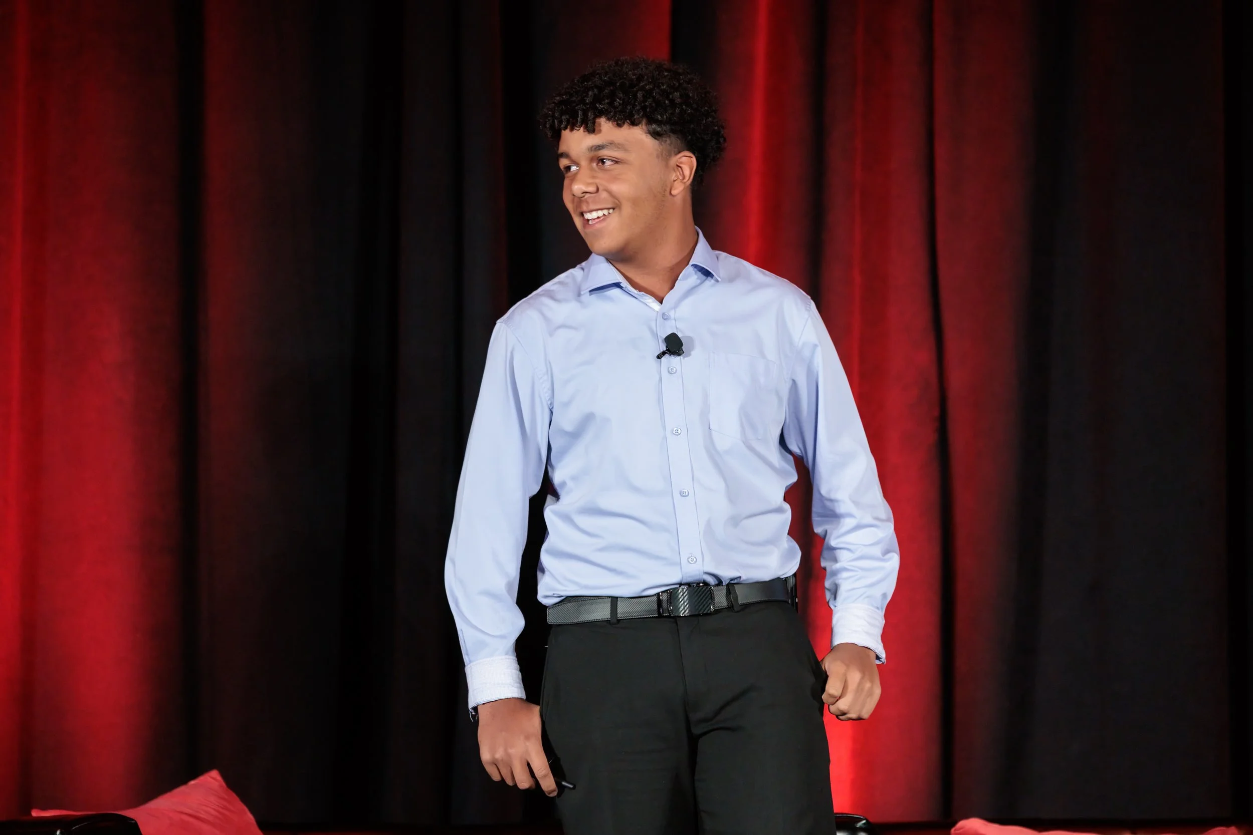 A young man in a light blue dress shirt and black pants standing in front of red and black curtains, smiling and looking to his right.