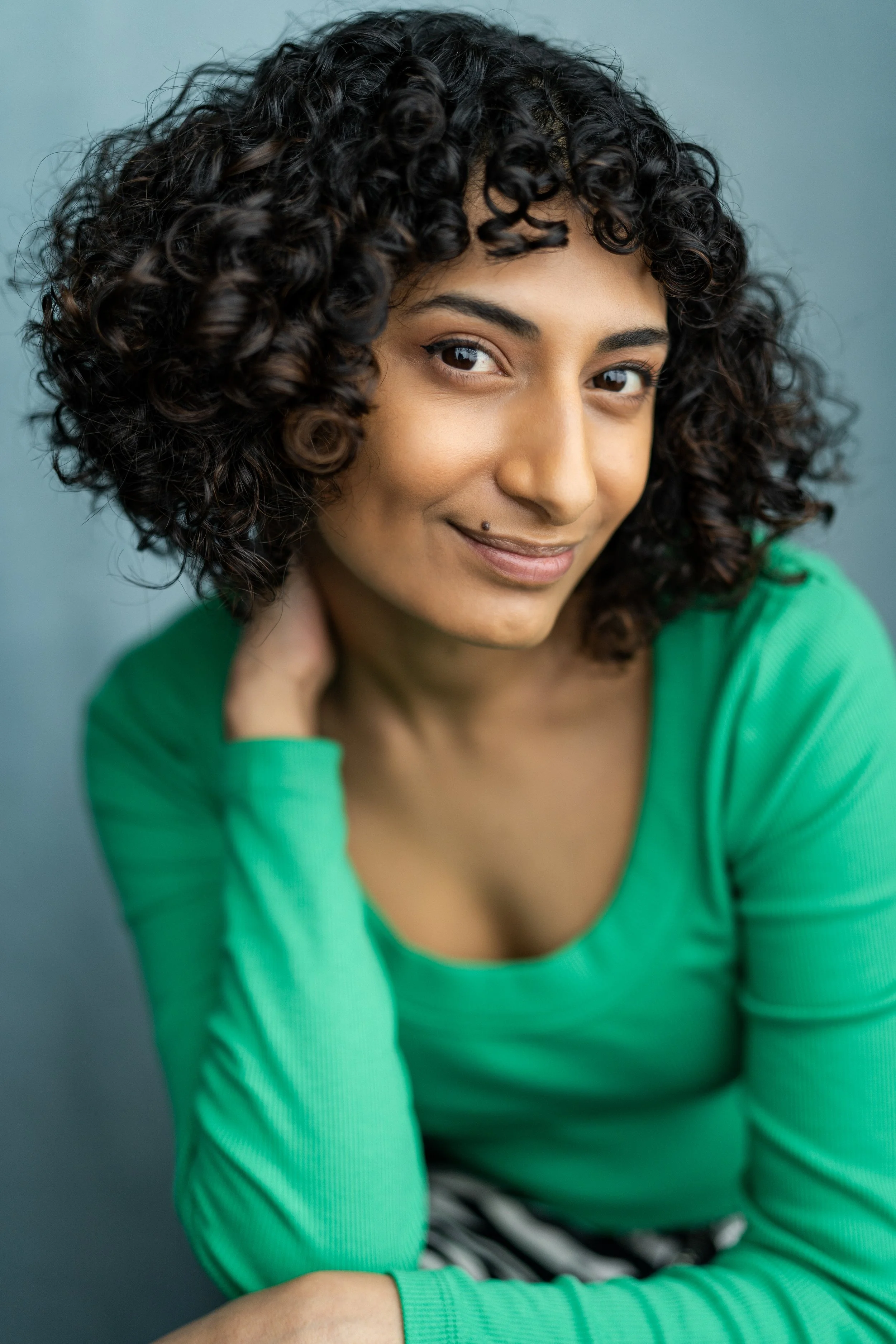 A young woman with medium skin tone, curly black hair, and wearing a green long-sleeve shirt, smiling gently at the camera, with one hand resting on her shoulder against a plain, gray background.
