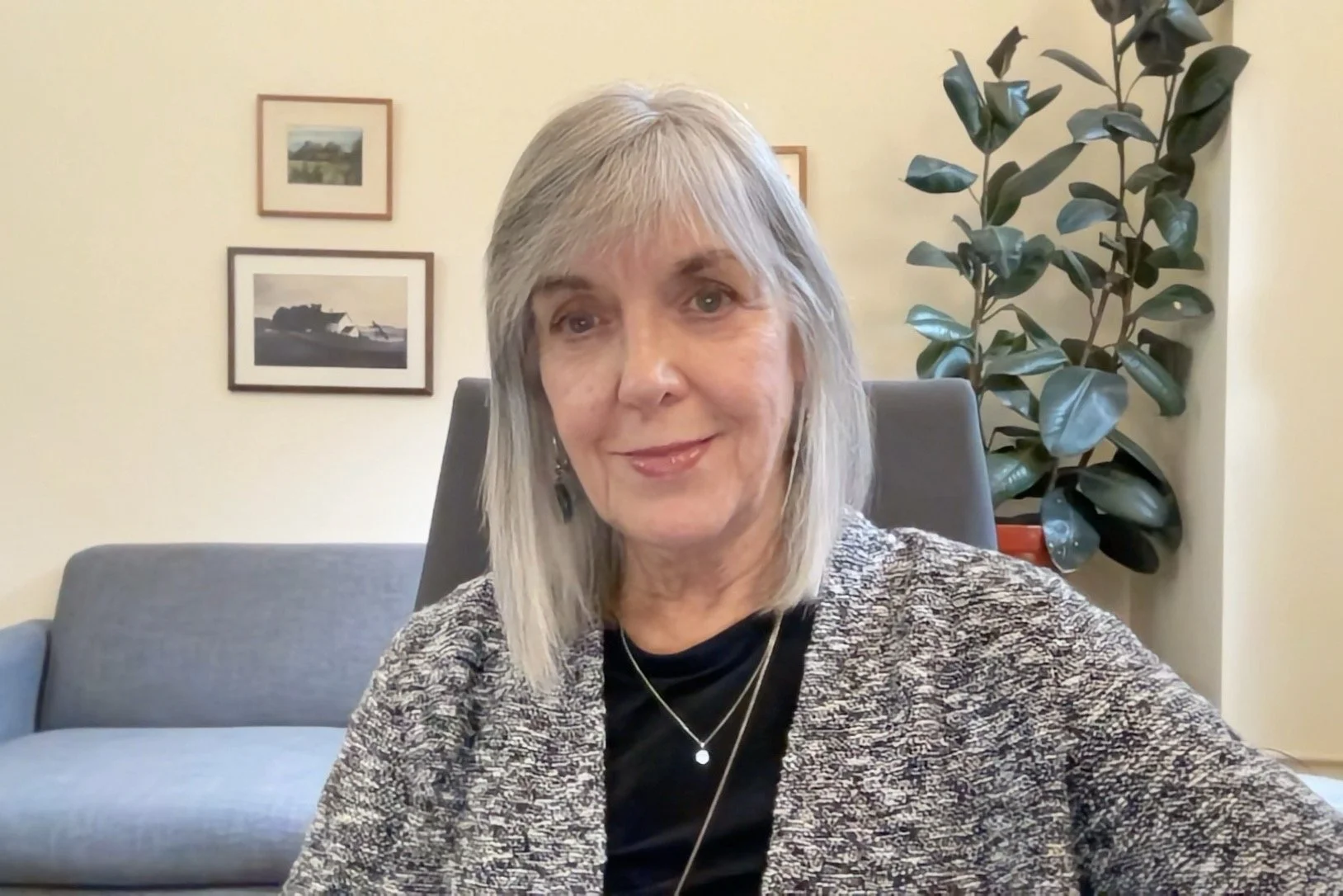 A woman with gray hair and light skin sitting in a home office or living room, smiling at the camera. Behind her, there is a blue sofa, framed pictures on a cream-colored wall, and a large potted plant with dark green leaves.