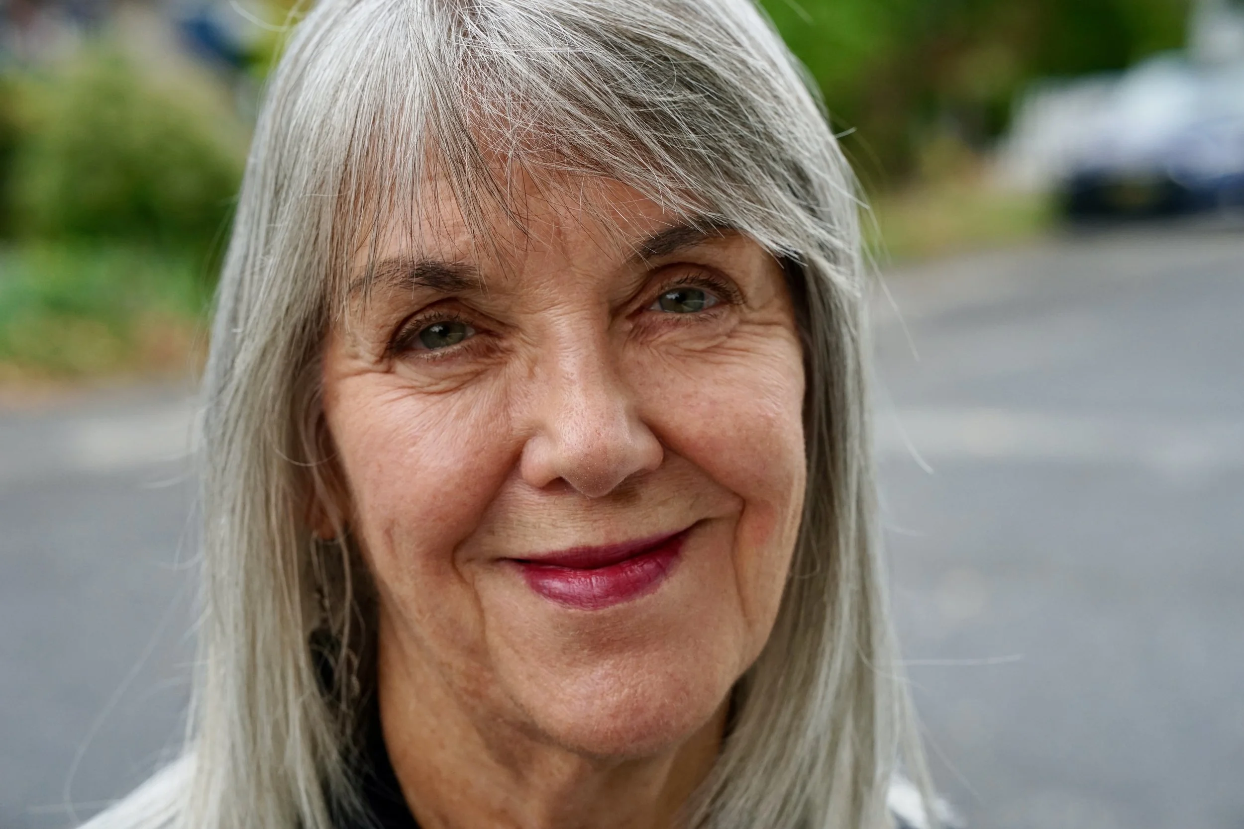 Close-up of a senior woman with long gray hair, smiling outdoors with blurred greenery and street in the background.