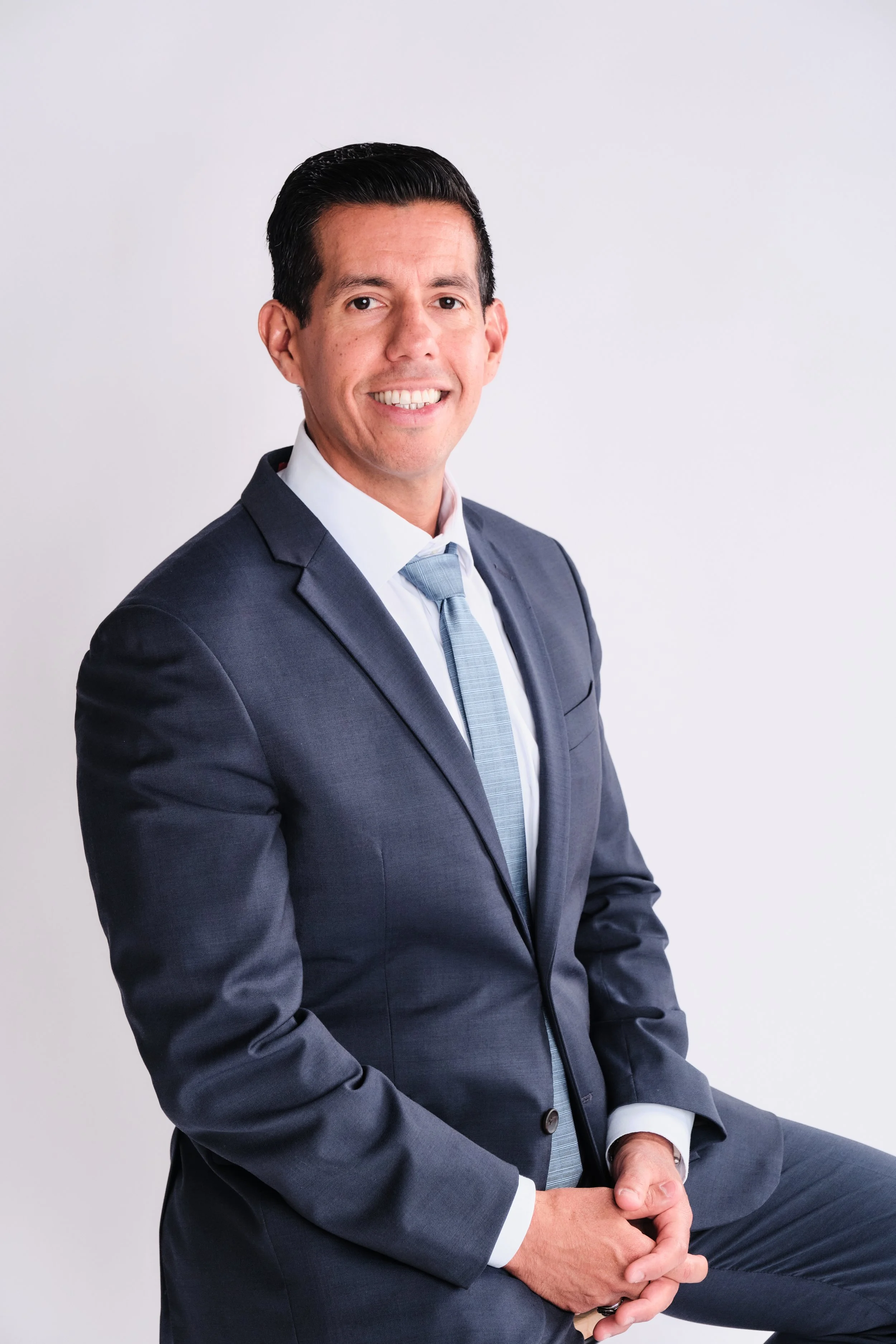 A man in a dark gray suit with a white shirt and light blue tie, sitting with his hands resting on his knee, smiling softly, against a plain light background.