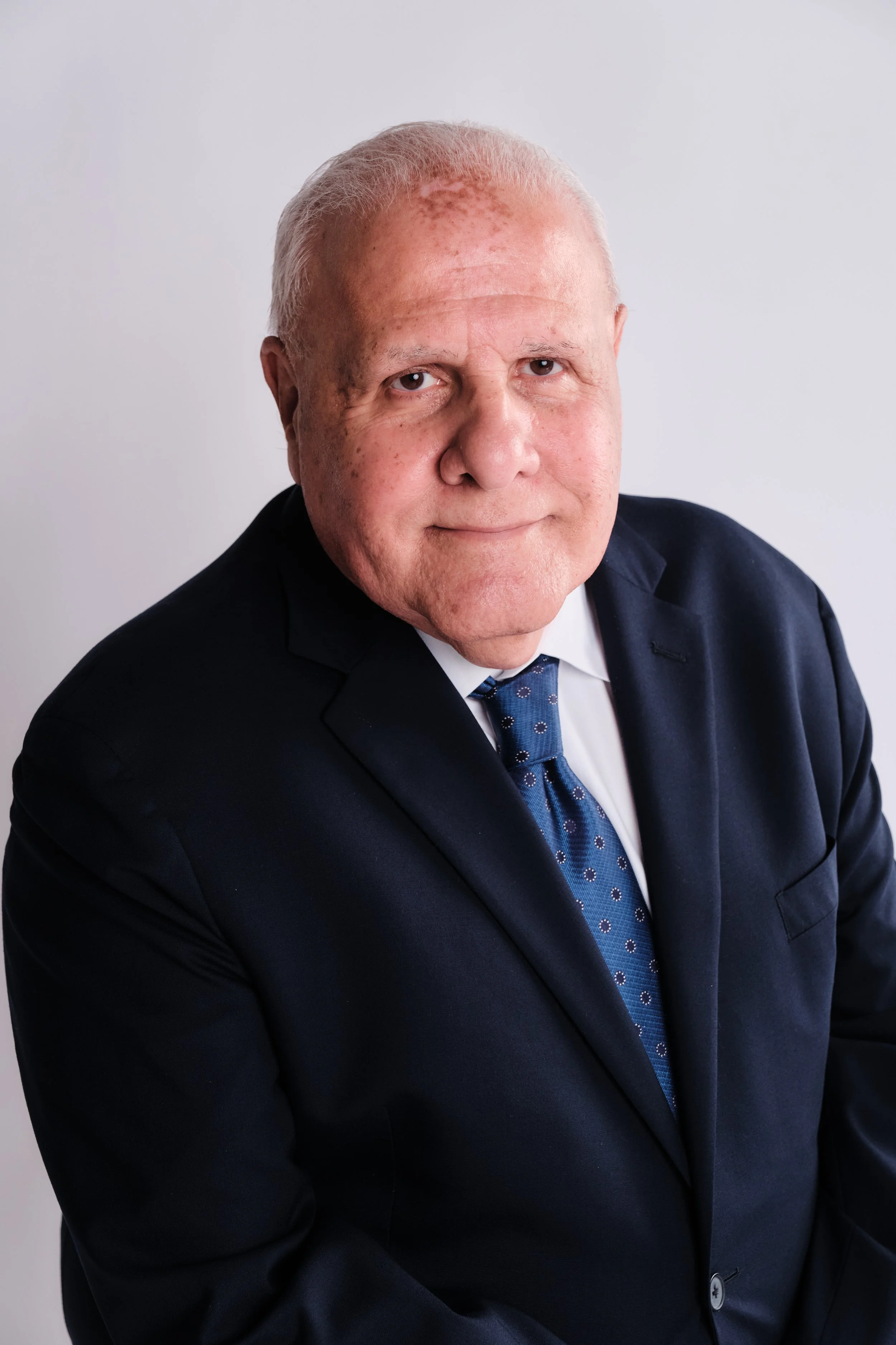 A middle-aged man dressed in a dark suit, white shirt, and blue patterned tie, posing against a plain light background, looking serious.