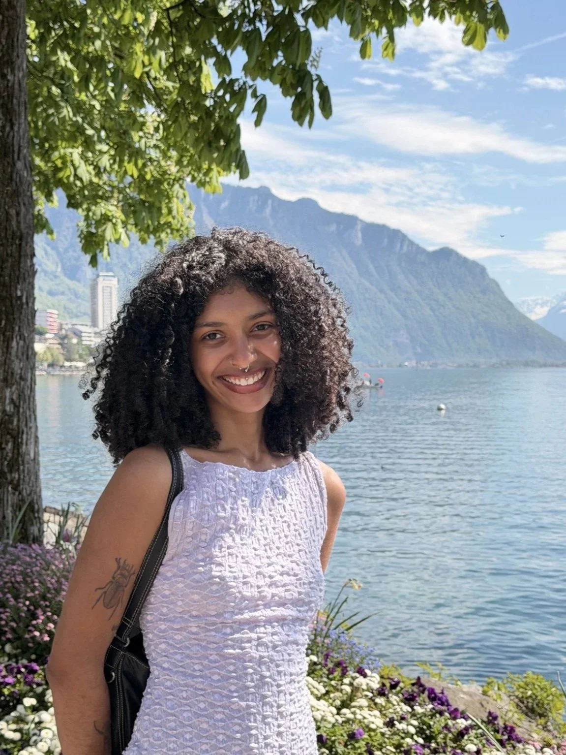Sorahya outdoors by a lake, with mountains in the background, trees overhead, and colorful flowers in the foreground.