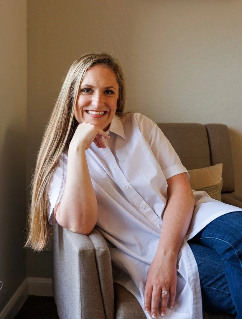 A smiling woman with long blonde hair, wearing a white shirt, sitting on a beige armchair in a room with beige walls.