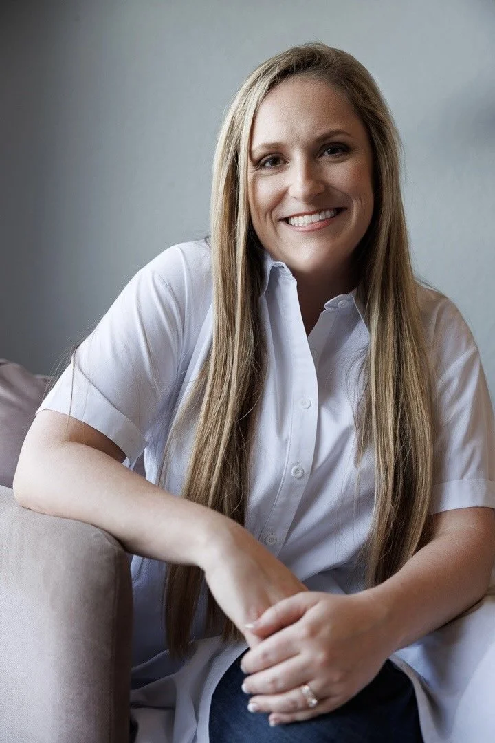 A woman with long blonde hair smiling while sitting on a beige couch, wearing a white button-up shirt