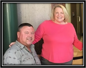 A man and a woman smiling, sitting inside a building near a green pipe and a door.