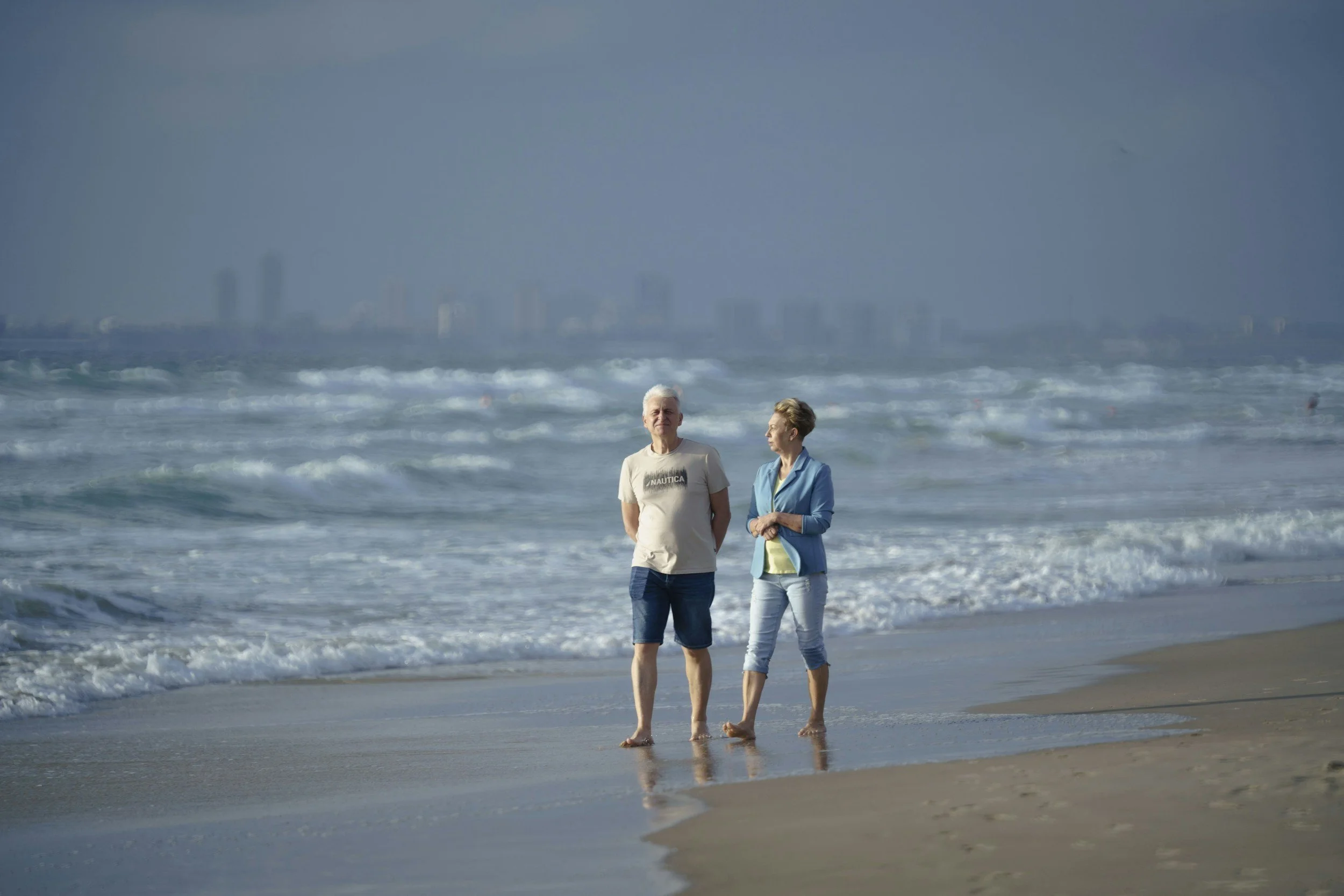 Two older adults, a man in a white t-shirt and shorts and a woman in a blue jacket and light pants, walking along the shoreline of a beach with waves and a city skyline in the background.