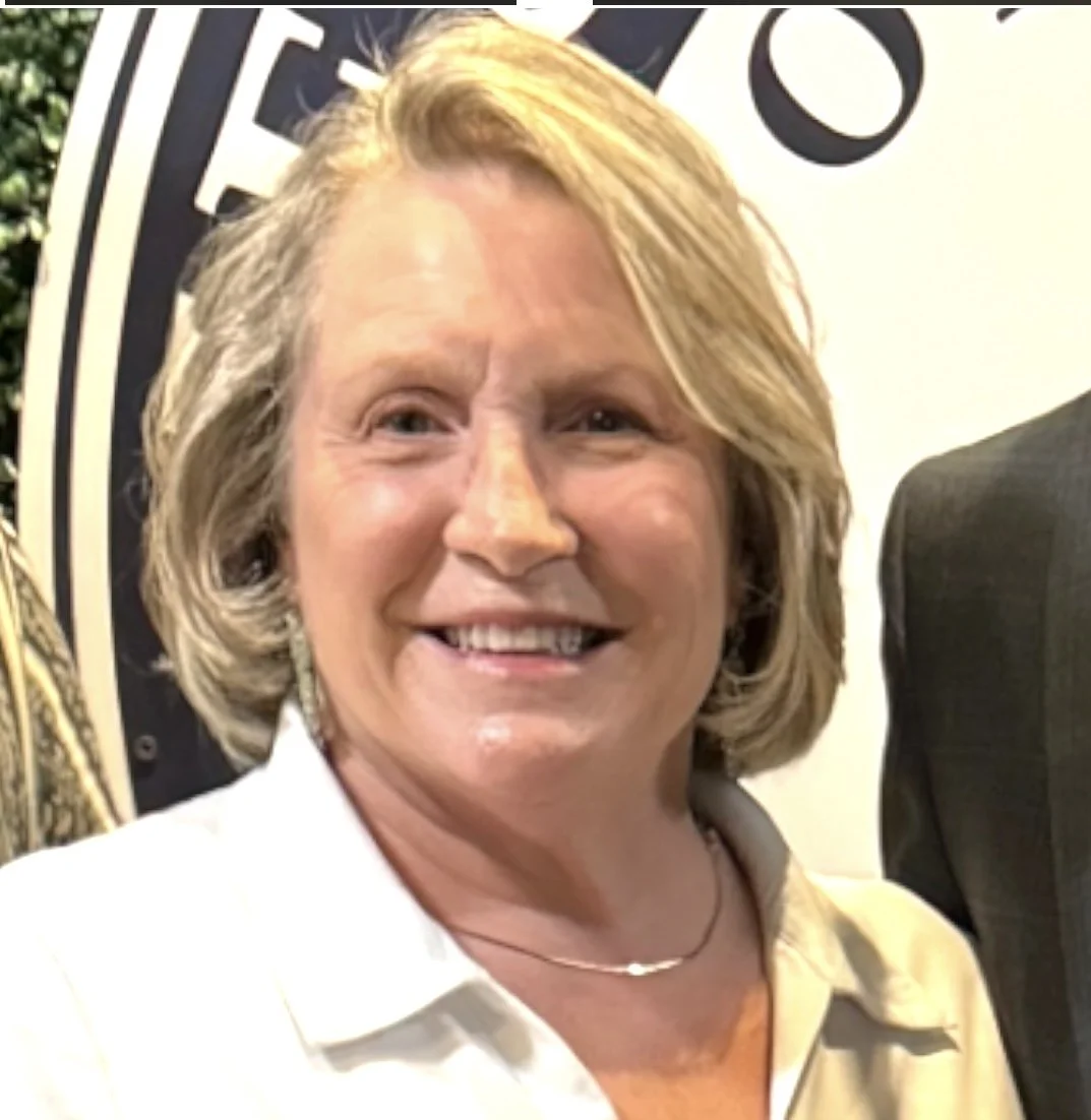 A woman with blonde, shoulder-length hair smiling, wearing a white shirt and a small necklace, standing in front of a sign with partially visible text.
