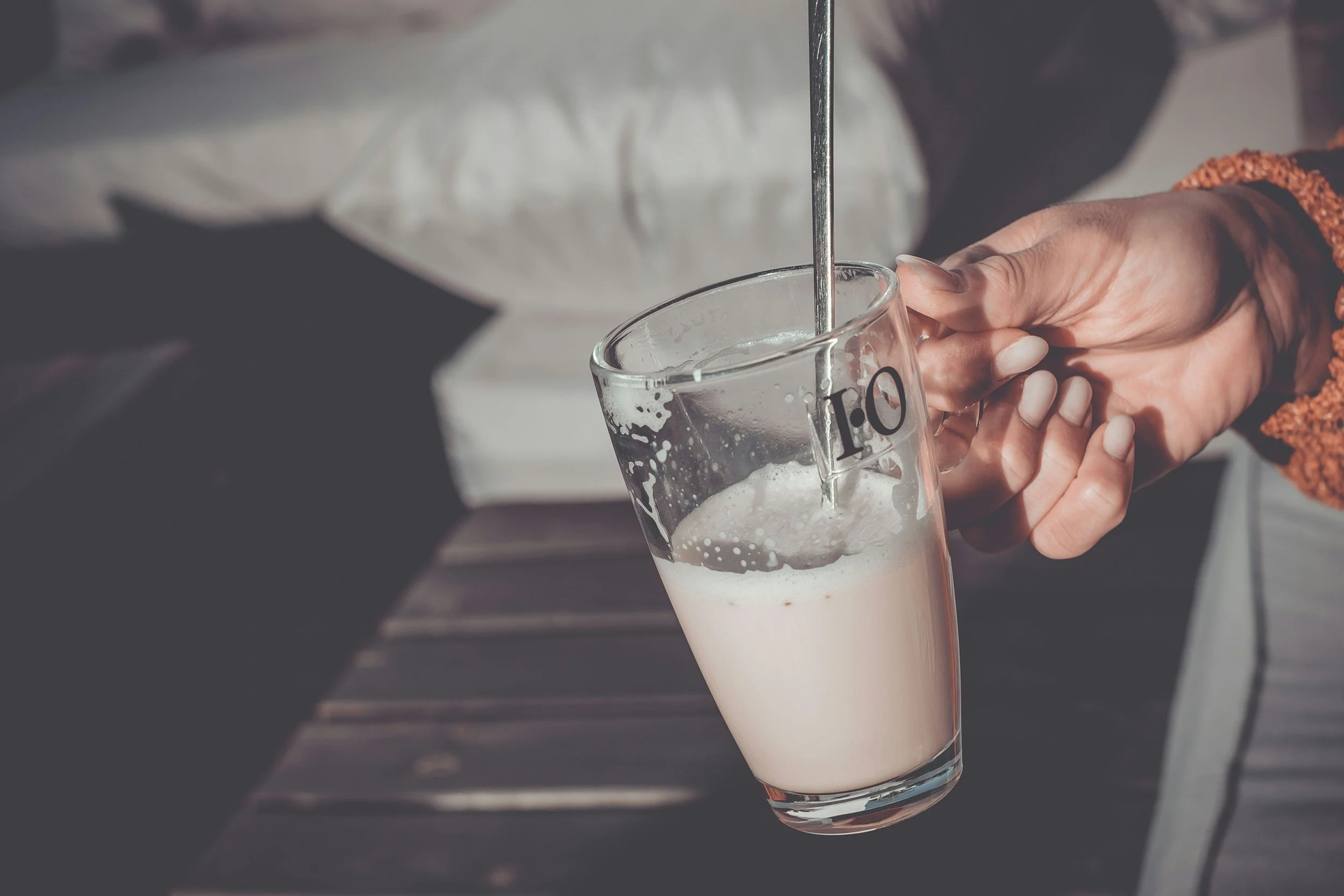 Person pouring a liquid from a metal spoon into a glass measuring cup filled with a white liquid, with the number 1.0 marked on the side.