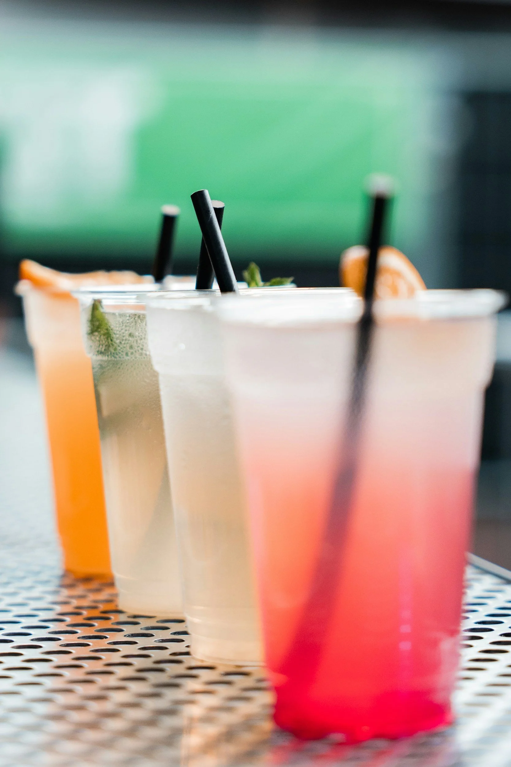Several colorful drinks with black straws on a perforated metal surface, against a blurred green and gray background.