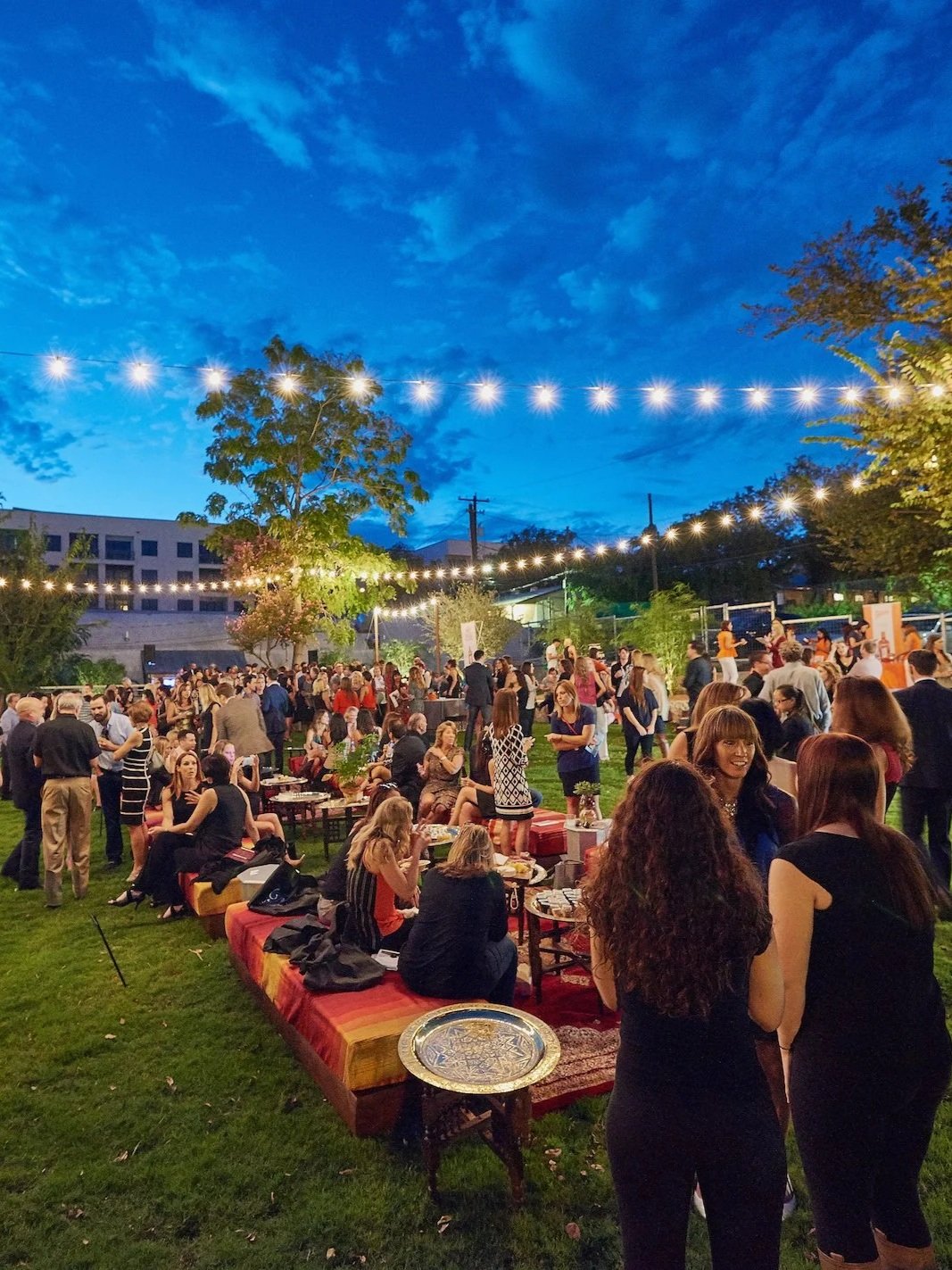 Outdoor evening gathering with many people socializing, sitting, and standing under string lights on a grassy area, with trees, a blue sky, and buildings in the background.