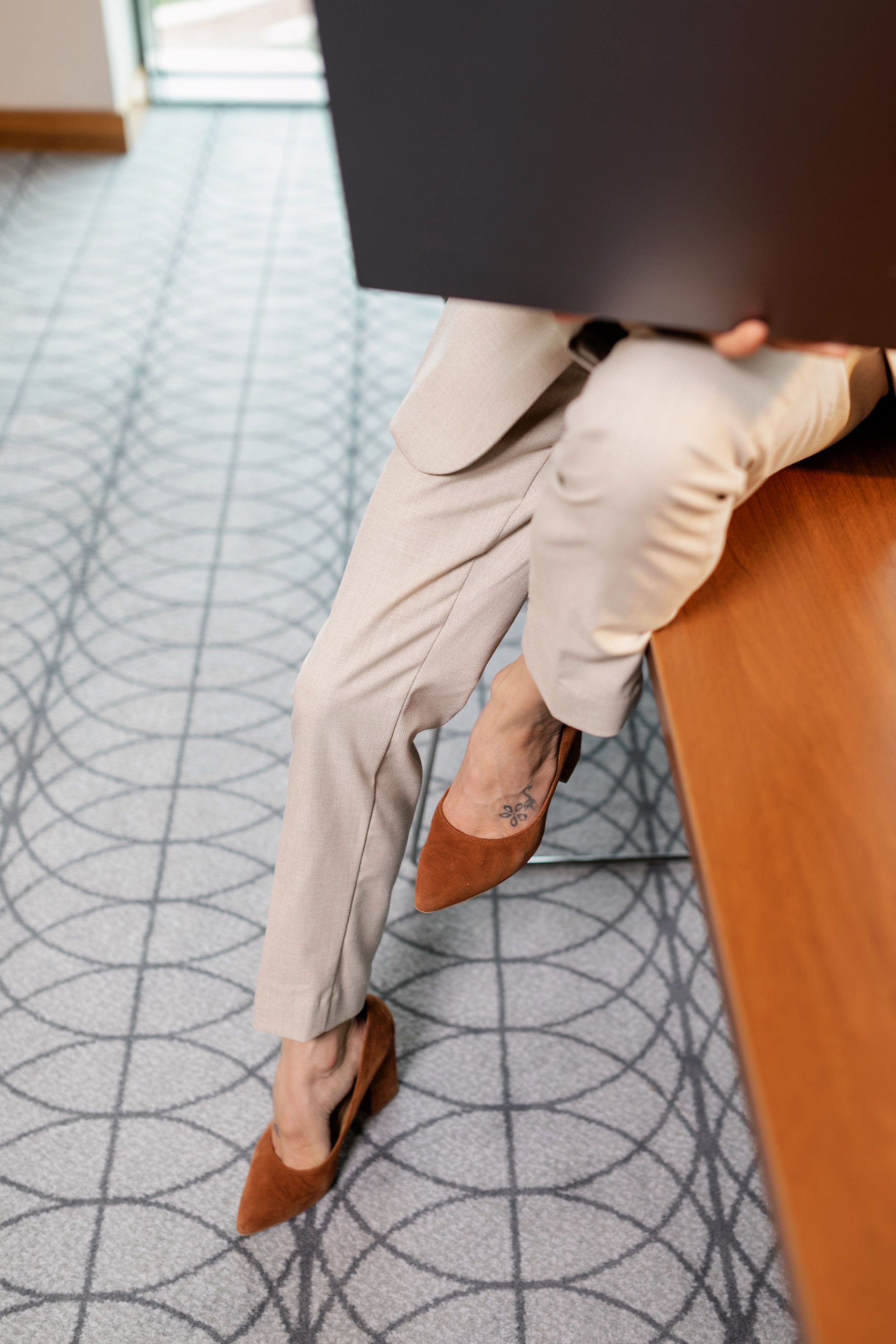 A woman seated at a wooden table wearing beige pants and brown high heels, with a tattoo on her ankle, holding a black folder.
