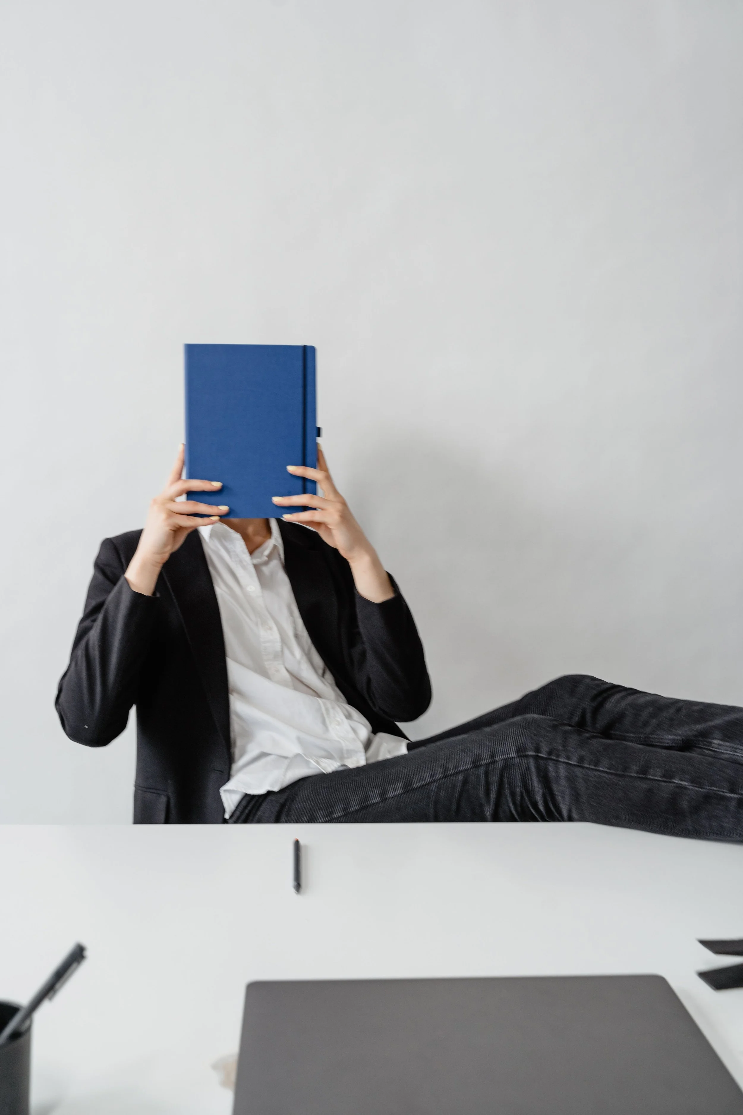 Person in business attire sitting at a white desk, holding a blue book or folder in front of their face. The person is wearing a dark suit and white shirt, with legs extended and crossed at the ankle. The background is plain light gray.