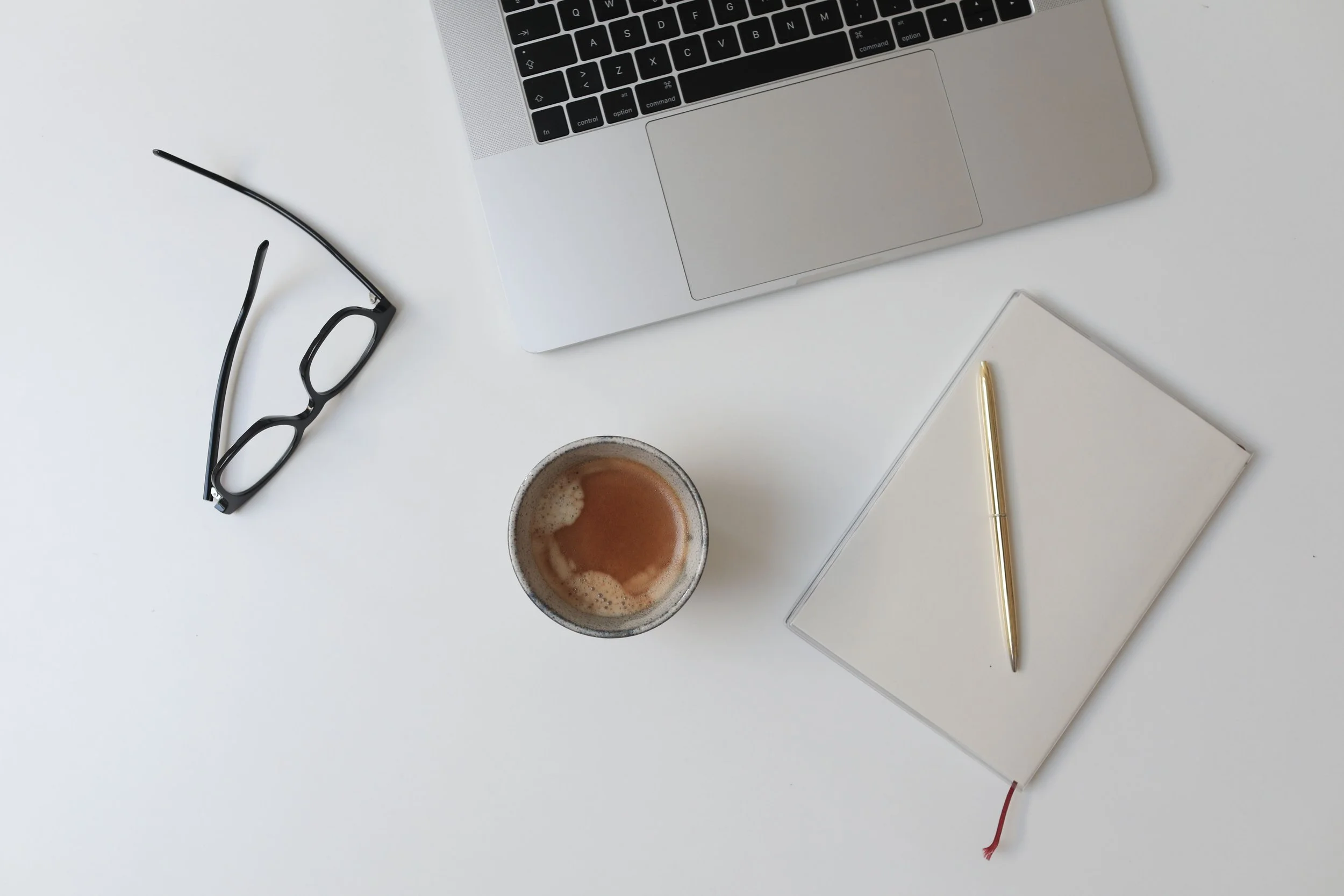 Top-down view of a workspace with a partial silver laptop, black glasses, a white notebook with a gold pen, and a cup of coffee on a white surface.