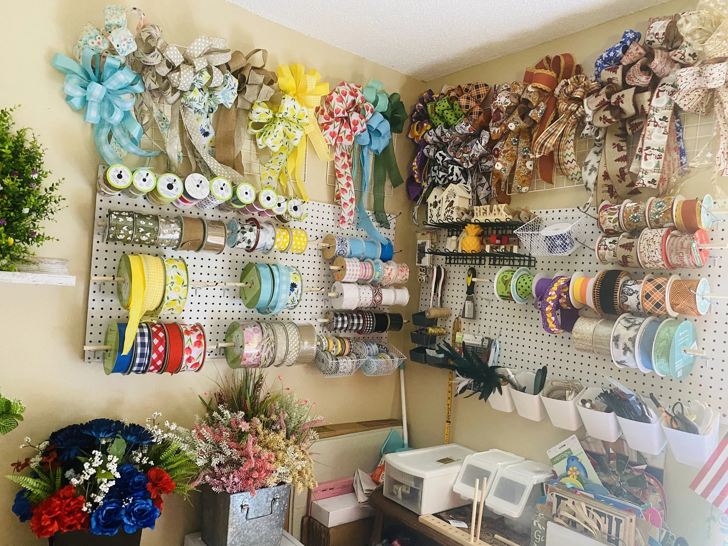 A wall display with various decorative ribbons tied into bows on hooks, colorful ribbon spools, foil and patterned paper rolls, and storage bins with craft supplies below. There are also floral arrangements with red, blue, pink, and white flowers at the bottom left corner of the image.