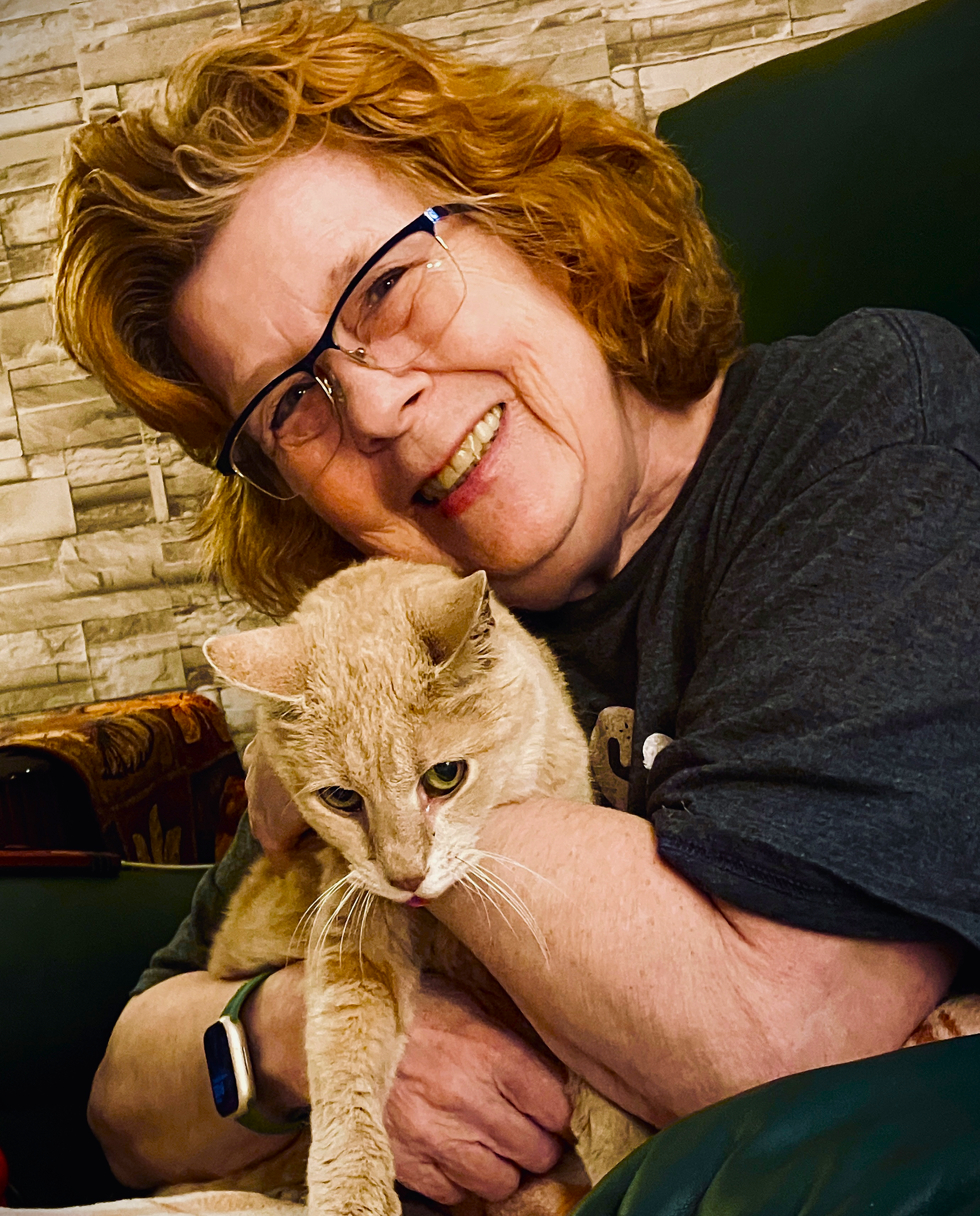 A woman with red hair and glasses smiling and hugging a ginger cat with green eyes.