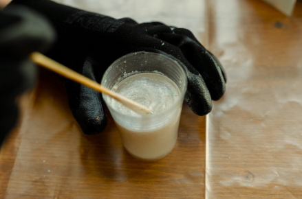 Person wearing black gloves stirring a glass of white liquid on a wooden surface.