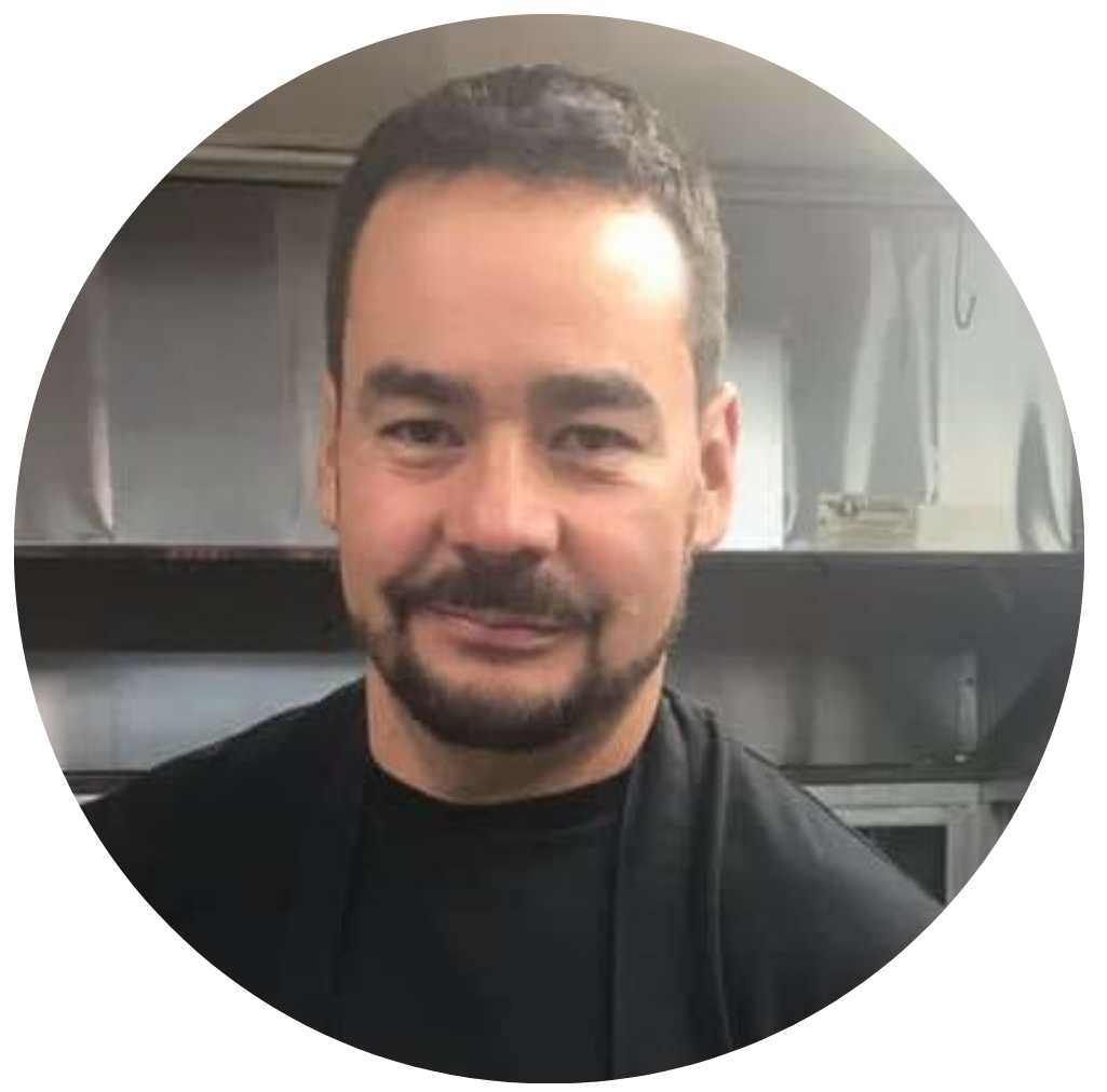 Hombre con barba y cabello oscuro, vestimenta negra, sonriendo, en un entorno de cocina con electrodomésticos de acero inoxidable.