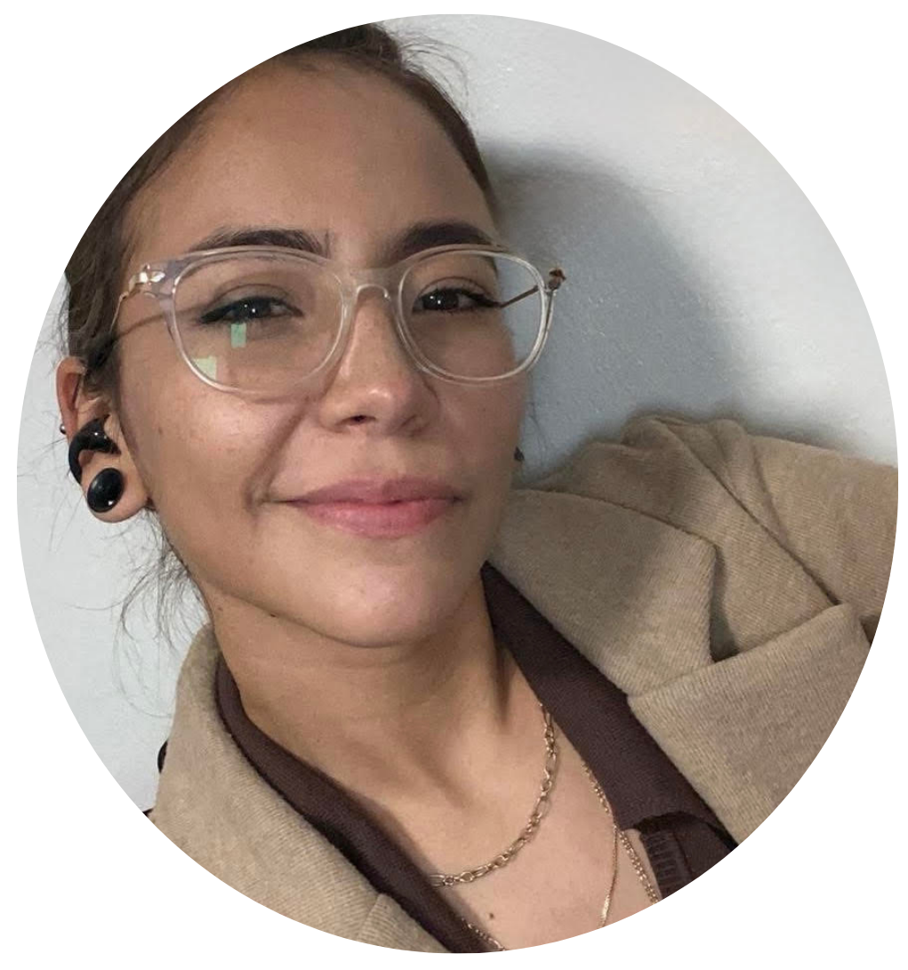 Retrato de una joven con gafas transparentes, pendientes grandes de caracol y un collar, sonriendo ligeramente frente a una pared blanca.