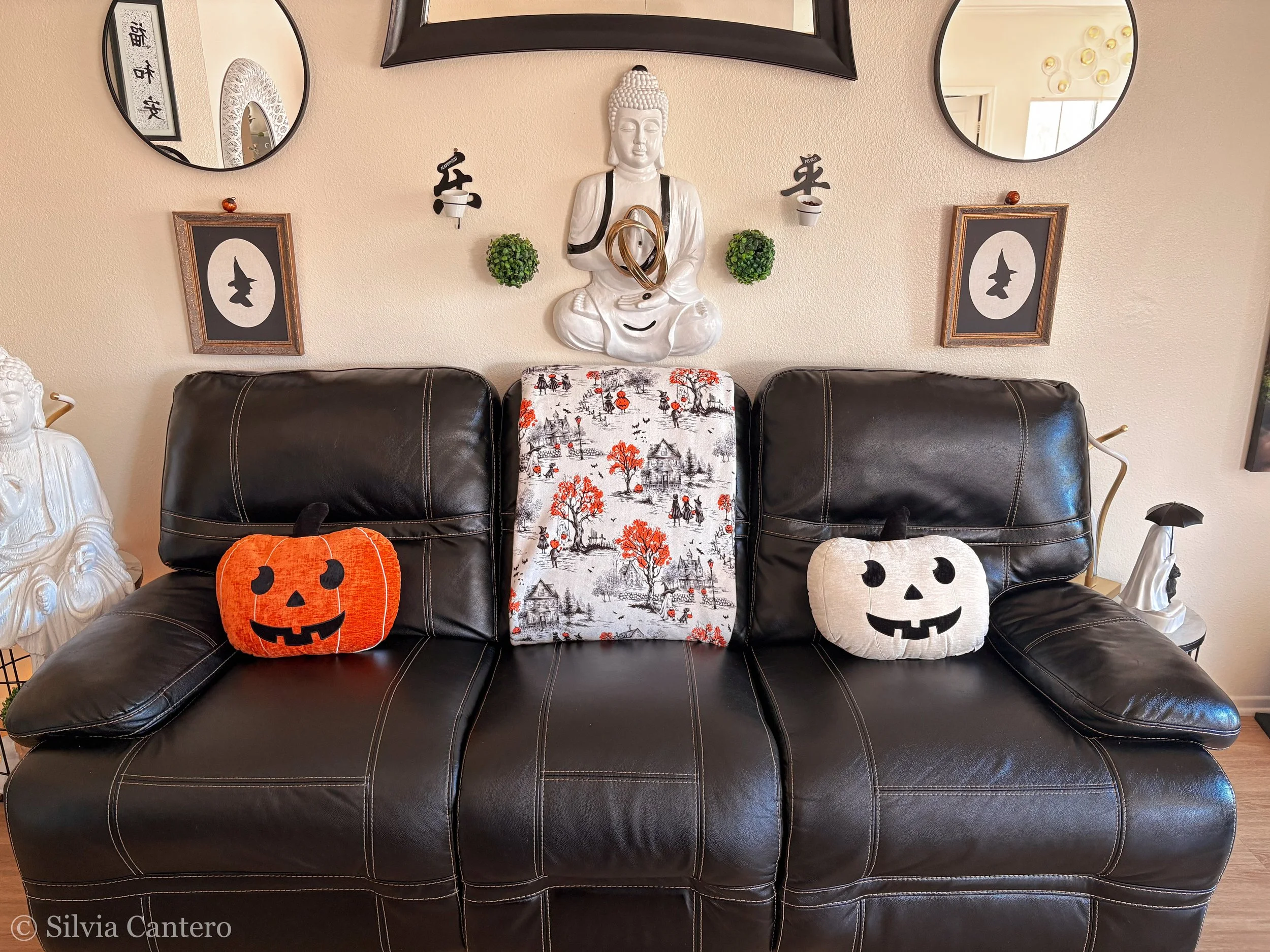 Living room with a black leather sofa decorated with Halloween-themed pillows, including a pumpkin and a ghost. On the wall behind the sofa are two mirrors, a white Buddha statue with vases and small greenery pots, framed artwork of a witch hat silho
