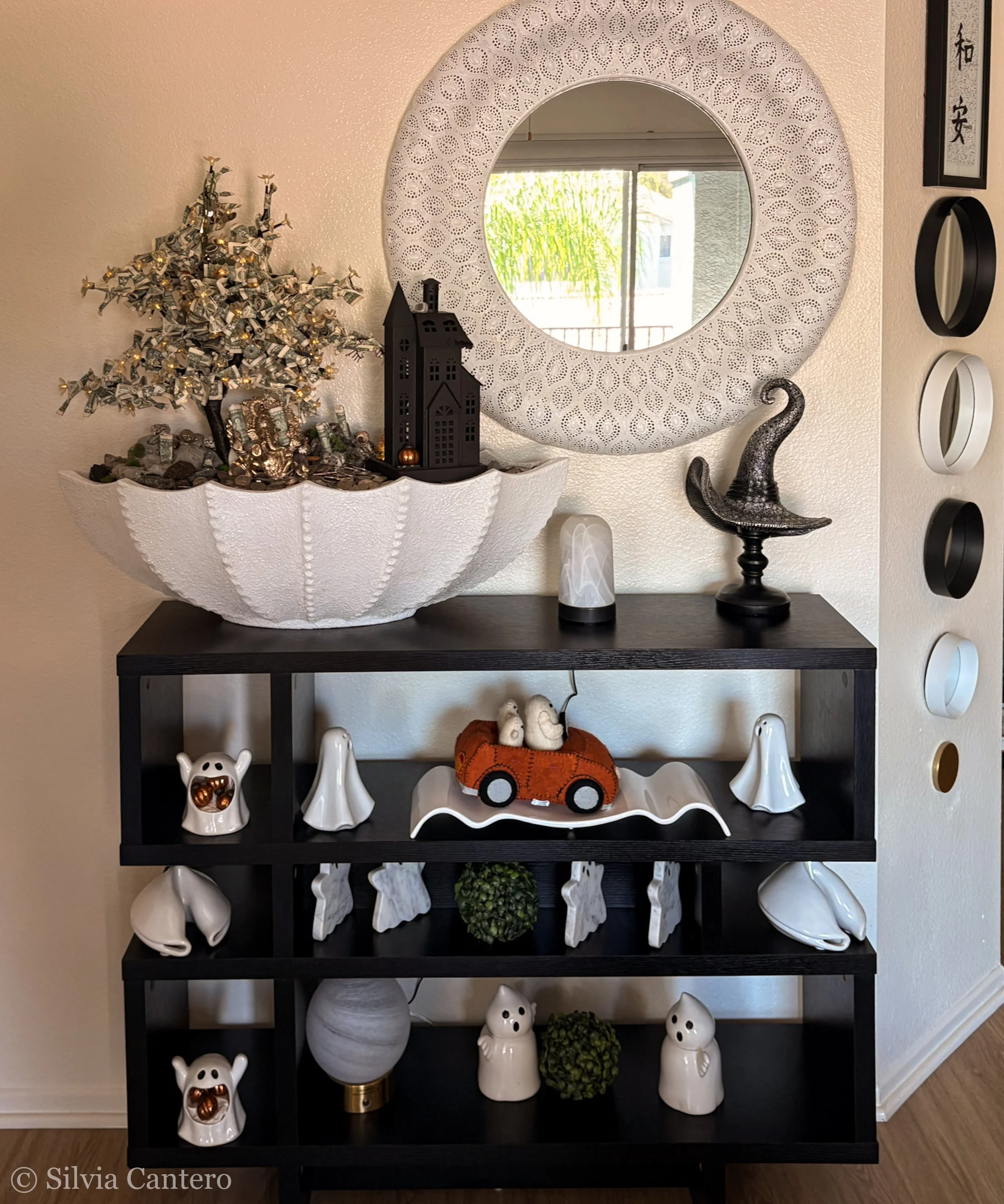 A black shelf with white ghost and skull decor and a small orange toy truck with skull decorations on top. Above the shelf is a large circular mirror with a white perforated frame. To the right of the mirror, there are vertical decorations with Asian