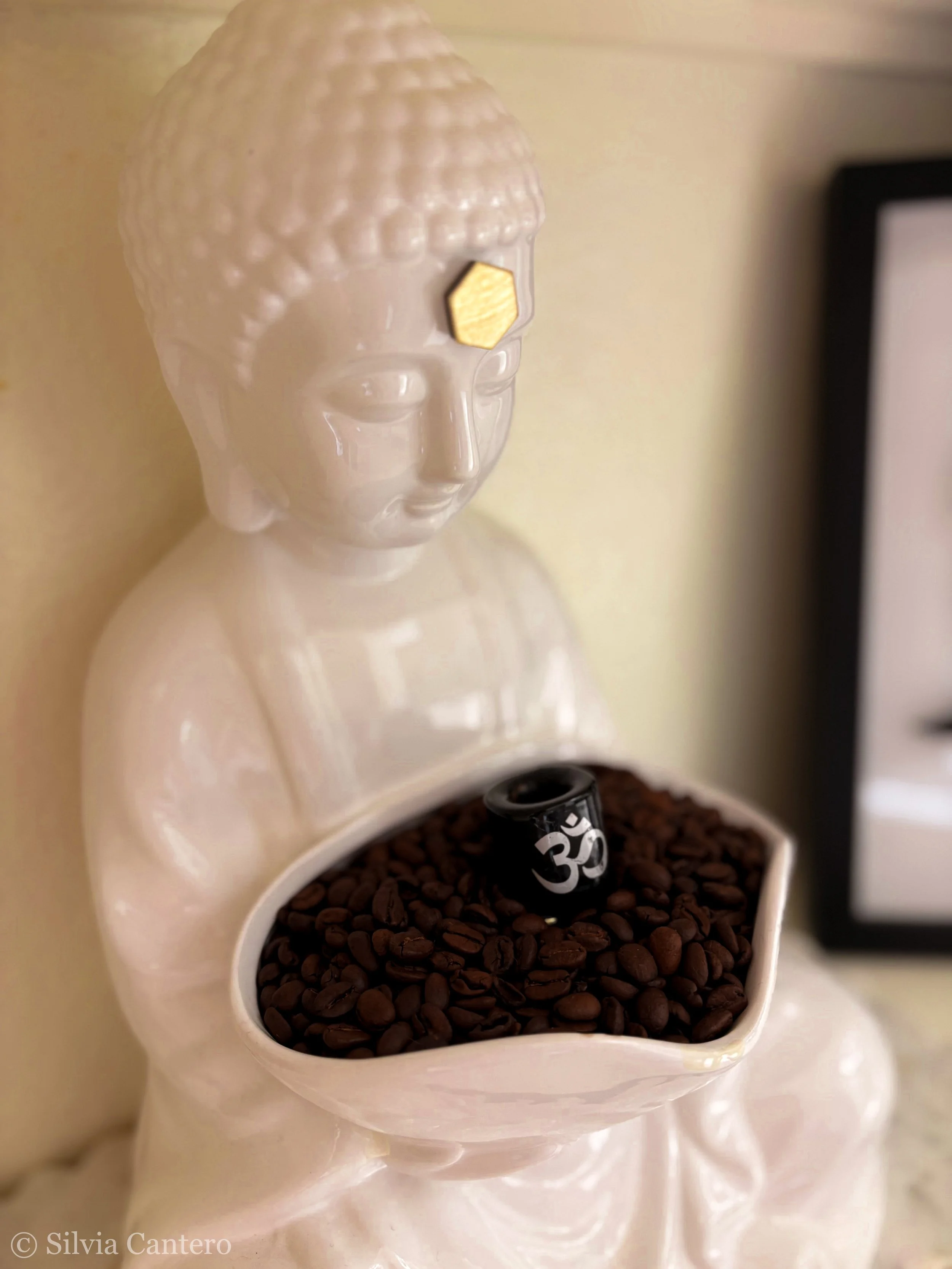 Close-up of two white ceramic bowls, one filled with coffee beans and the other with a black Om symbol engraved on a small, black object, possibly a spoon. There is a small, golden hexagon-shaped item attached to the bowl with the coffee beans. The b