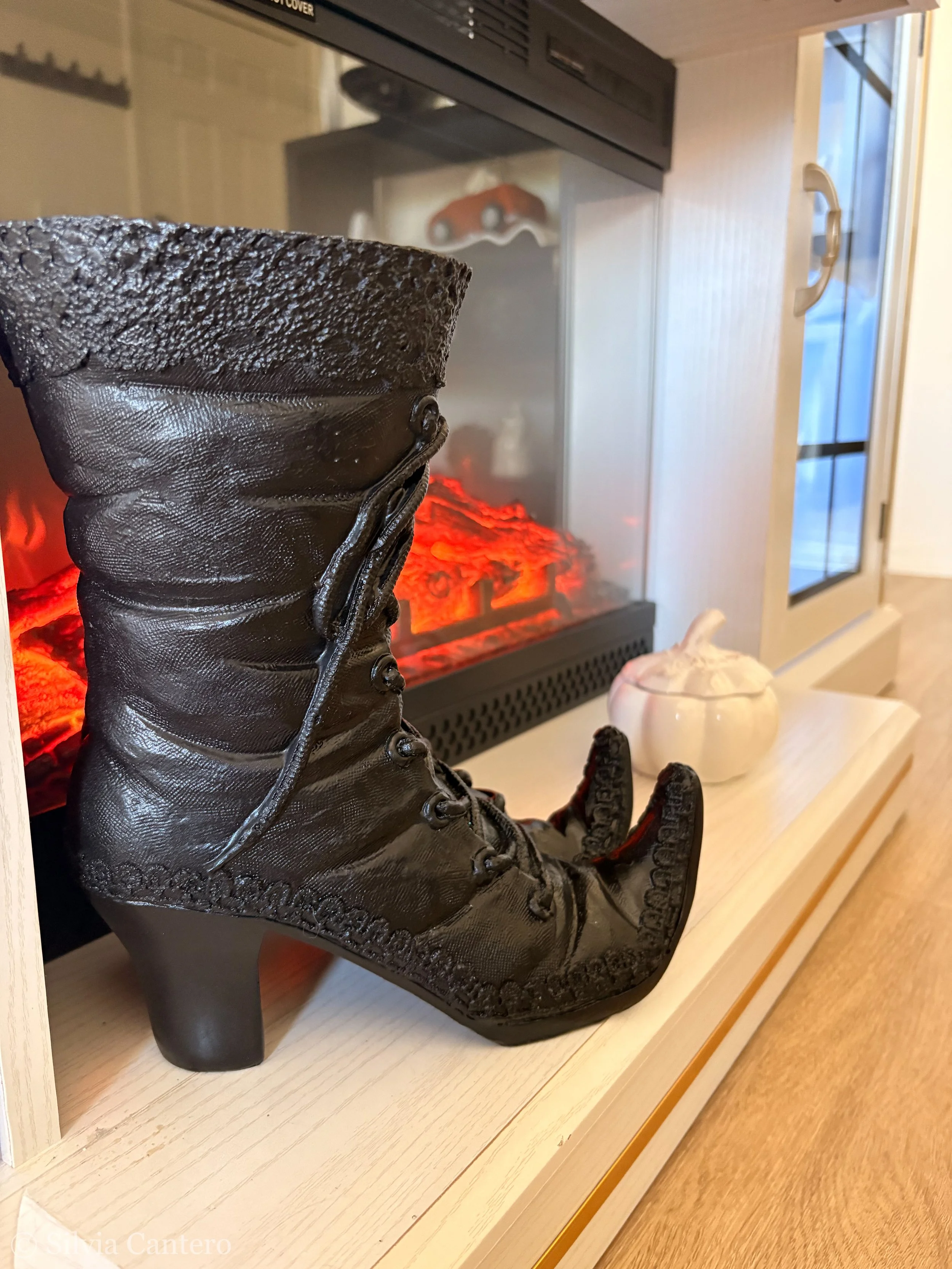 A black high-heeled boot sculpture with a textured surface, placed on a wooden display shelf in front of a fireplace with glowing red embers.