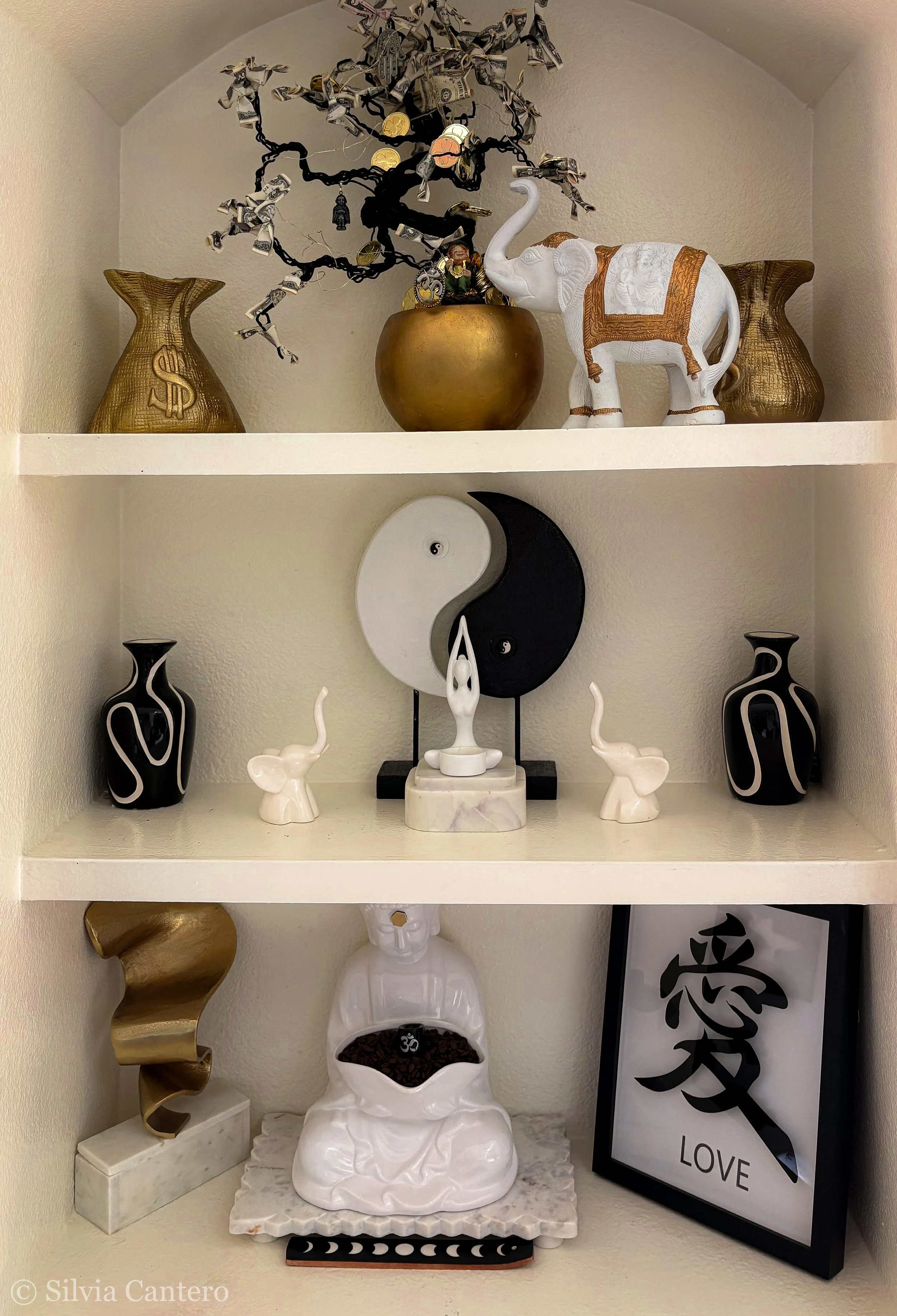Decorative shelves with various objects including golden vases, a paper money tree, a white elephant figurine, black and white vases, small elephant figurines, a white Buddha statue with a bowl of coffee beans, and a framed art piece with Chinese cha