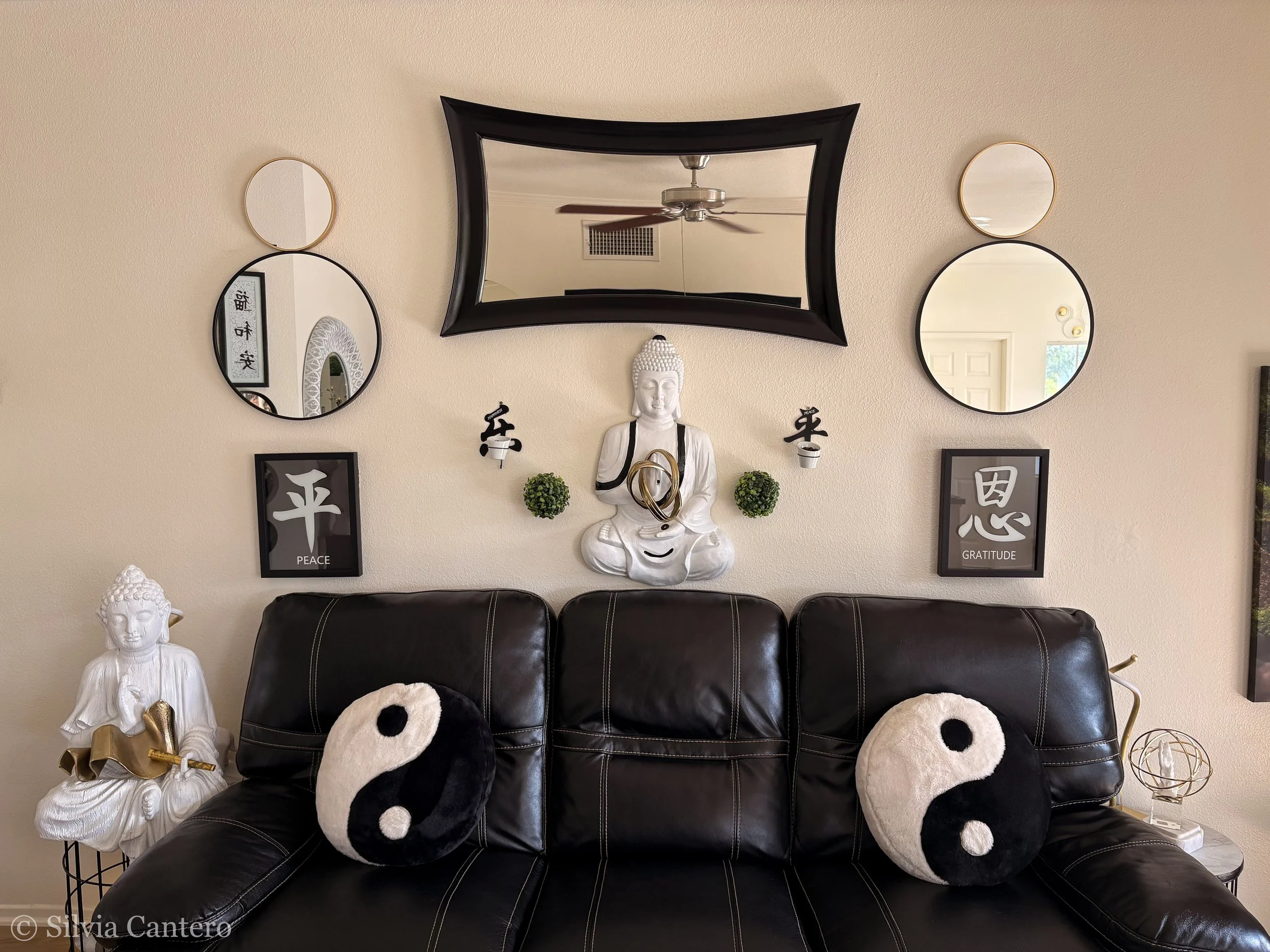Living room wall decor including a large black-framed mirror, five round mirrors of varying sizes with gold and black frames, two black-and-white panda plush pillows on a black leather sofa, a white Buddha statue with a golden ornament, and two frame