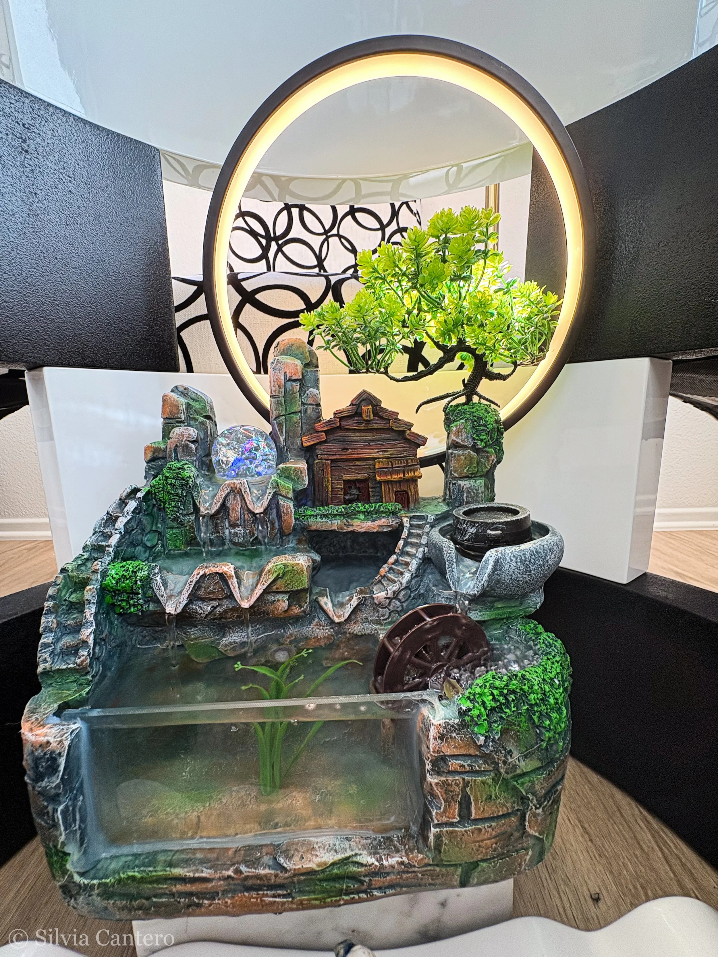 An indoor decorative water fountain with small waterfalls, a miniature house, and green plants, illuminated by an LED ring light behind it.
