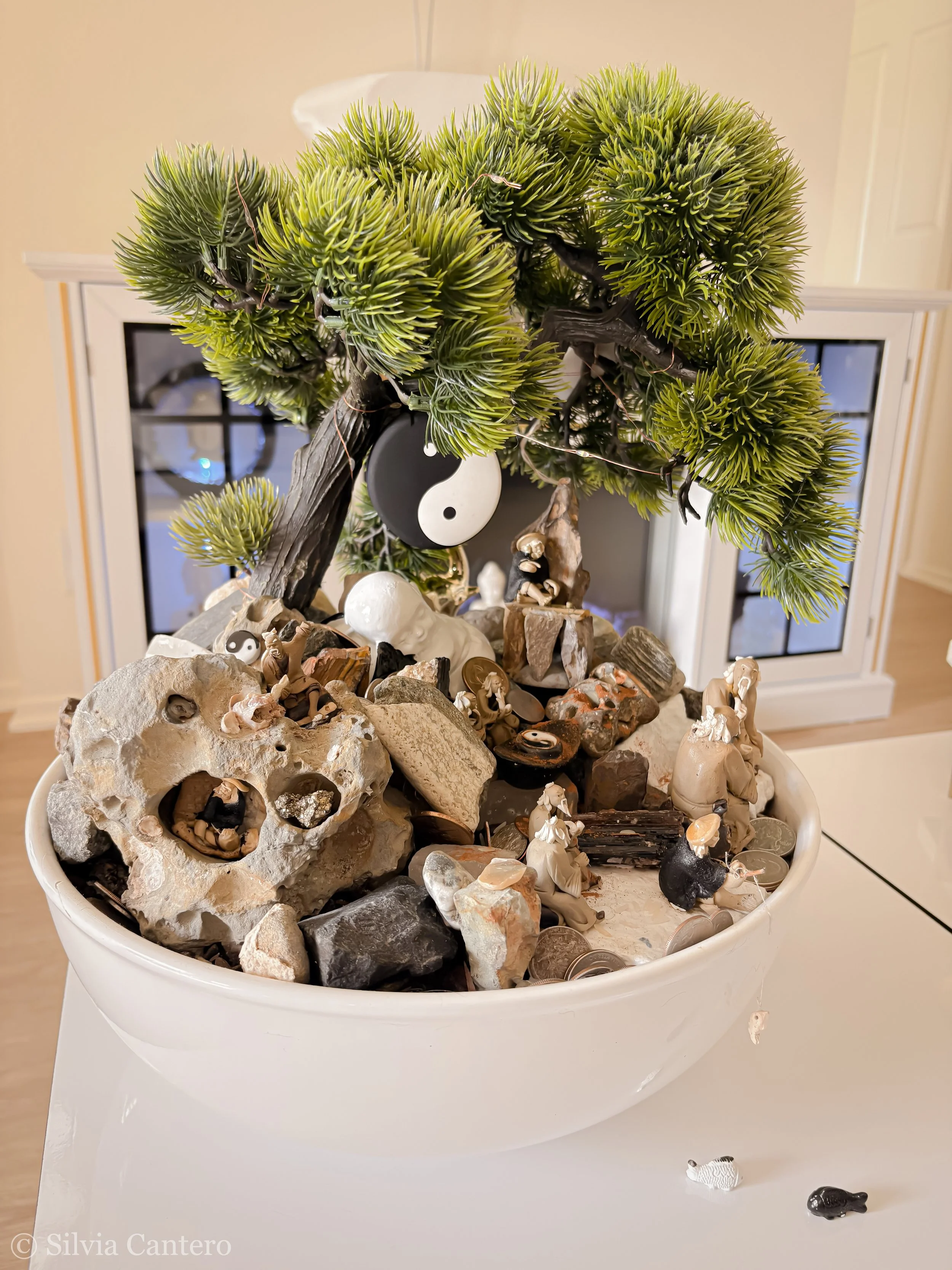 A white round container filled with rocks, small figurines, and decorative items, topped with a small bonsai tree and hanging yin-yang symbols.
