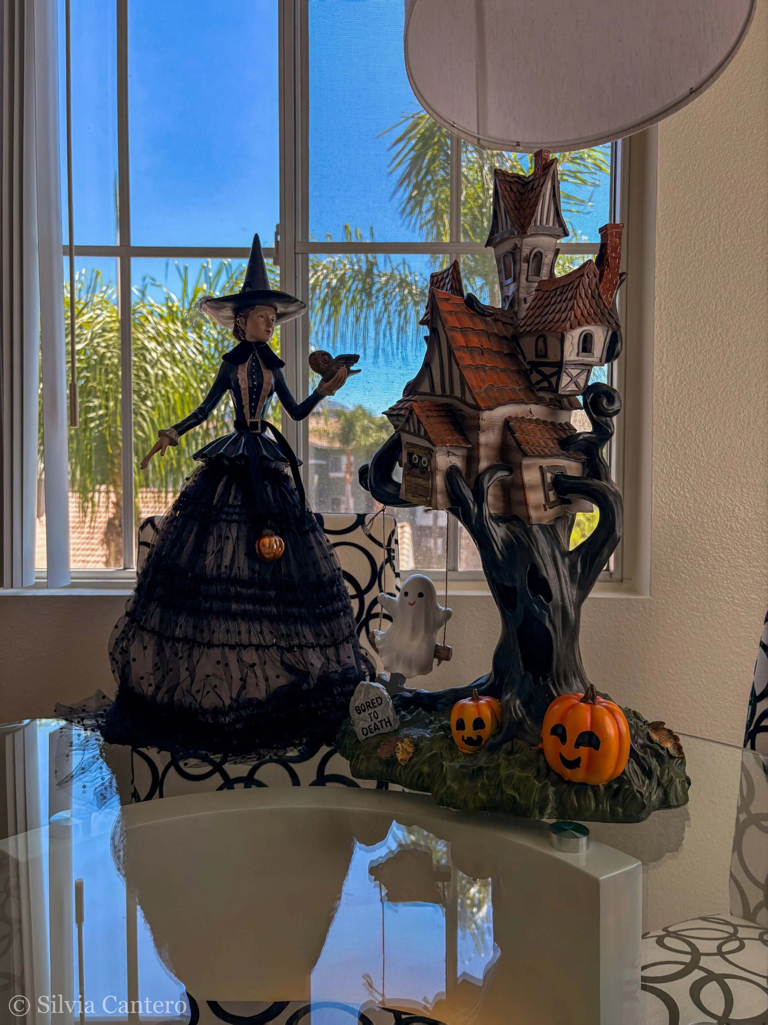 Halloween decorations on a table near a window featuring a witch figurine, haunted house, ghost figure, small pumpkins, and a sign that reads 'BORED TO DEATH'.