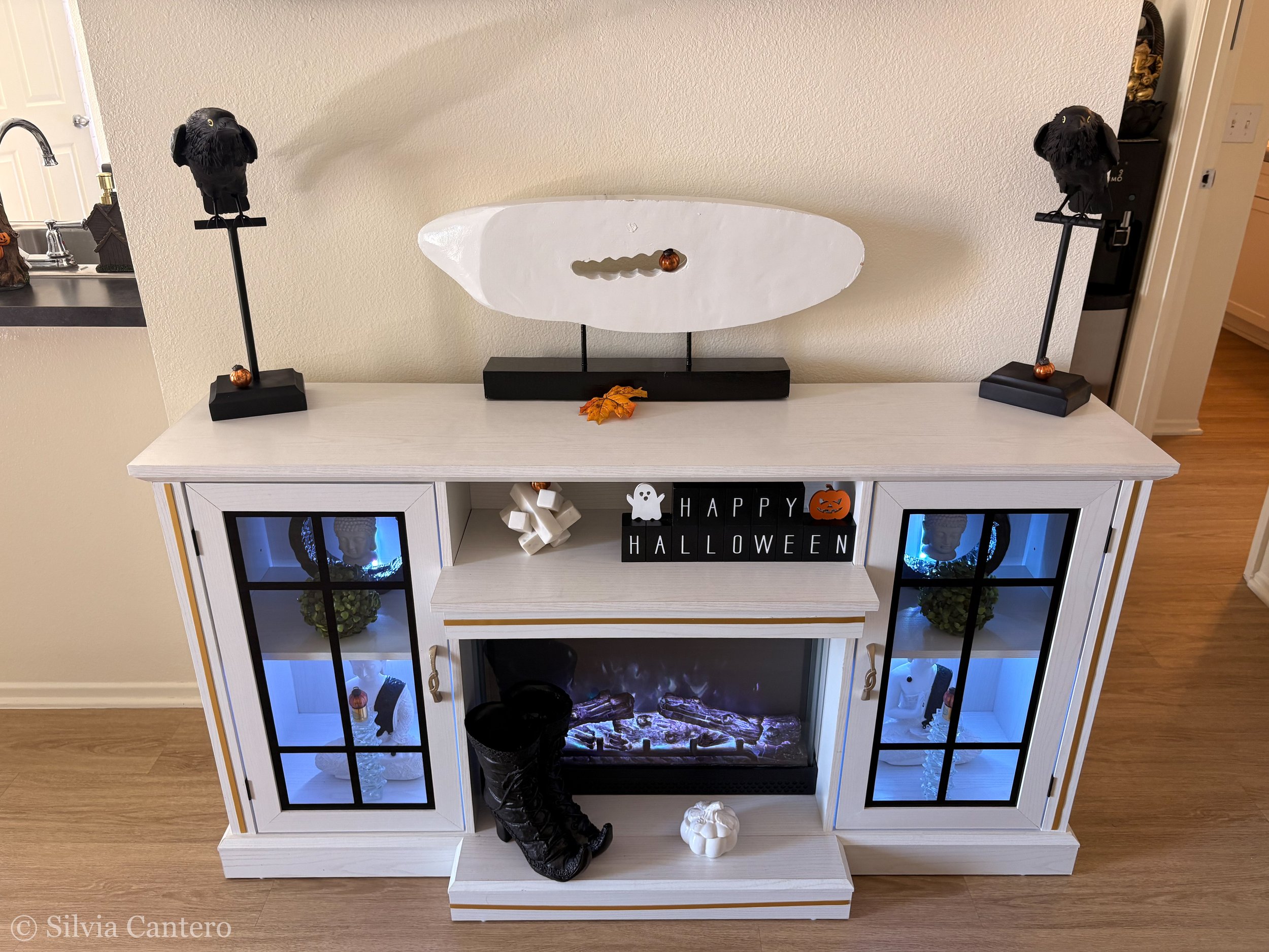 A Halloween-themed decoration display on a white cabinet with glass-paneled doors. The display includes a pair of black crow figurines on tall stands, a white decorative object with a pumpkin, a 'Happy Halloween' sign with a ghost and pumpkin, and a 